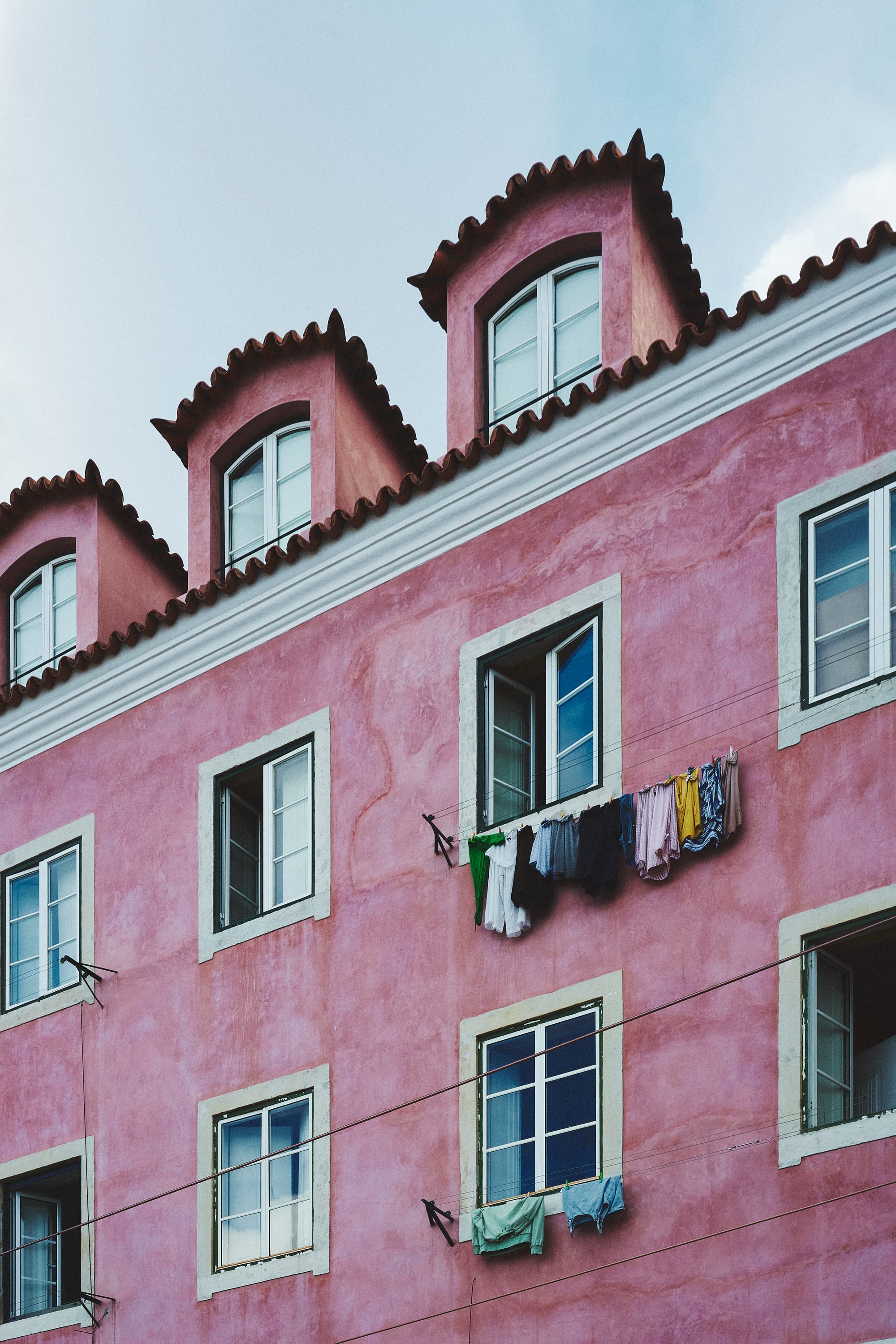 A pink building facade in Lisbon with laundry hanging from an open window between traditional tiled dormer windows