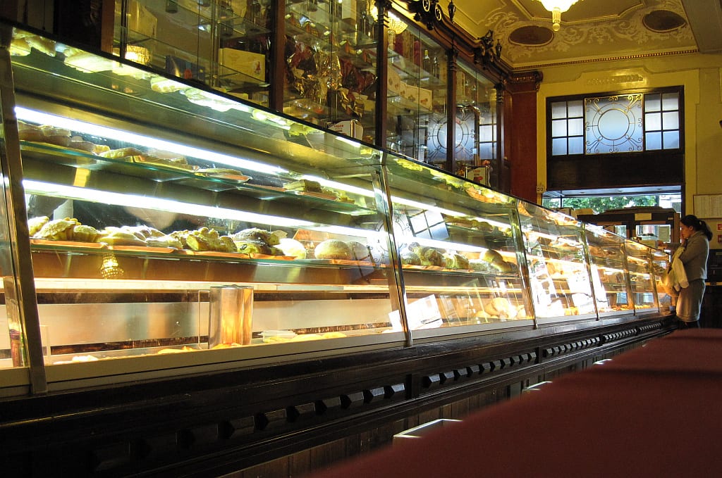 A long glass pastry counter filled with rows of cakes and bread inside an ornate old café, with a customer standing at the far end