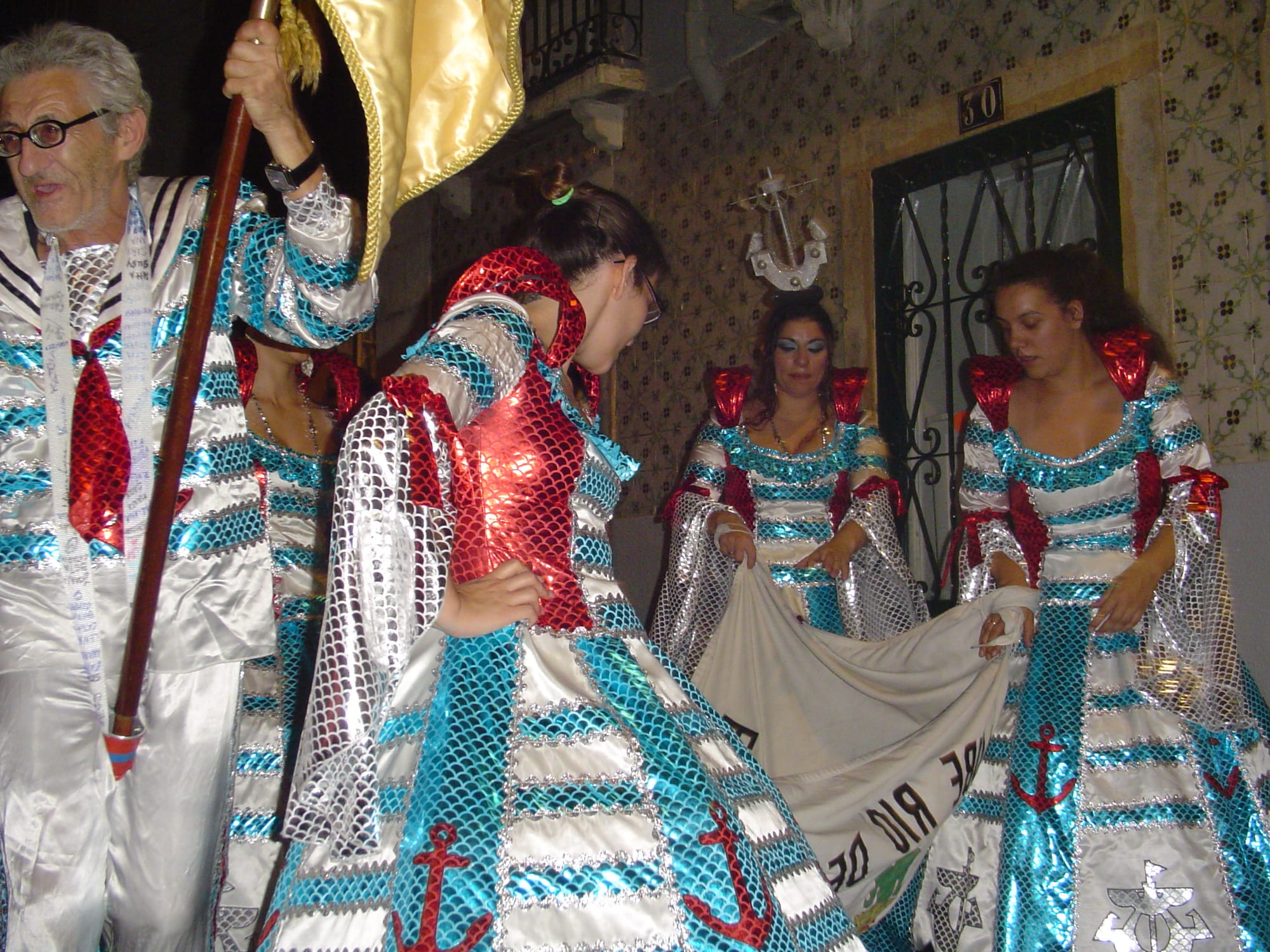 Marchers in shimmering costumes decorated with anchors and fish-scale patterns in turquoise, red and silver, one man holding a flag on a pole, gathered in a narrow street beside a tiled wall at night