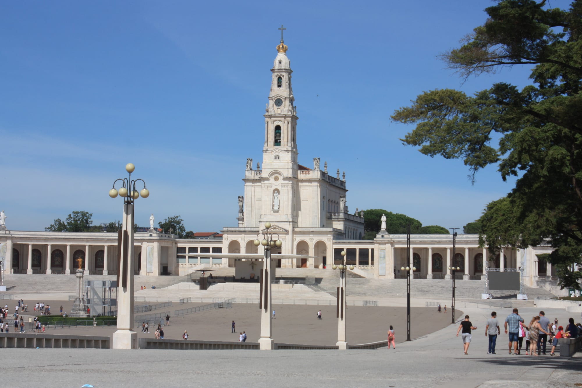 A white basilica with a tall bell tower topped by a crown and cross, flanked by curving colonnades, overlooking a wide paved esplanade with lamp posts and a few scattered visitors under a blue sky