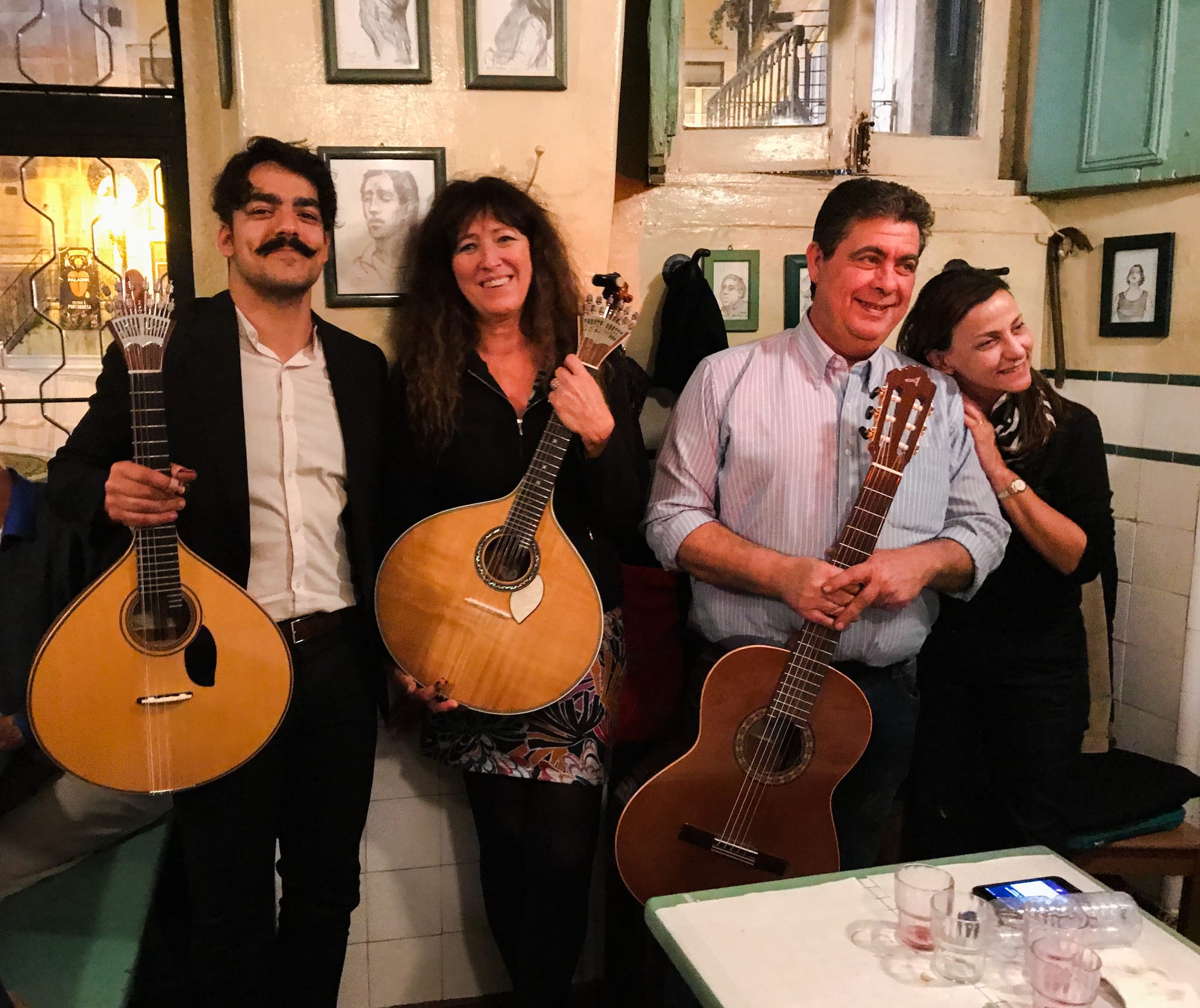 Four people standing in a small tiled tavern with framed drawings on the wall, two of them holding pear-shaped Portuguese guitars and one holding a classical guitar