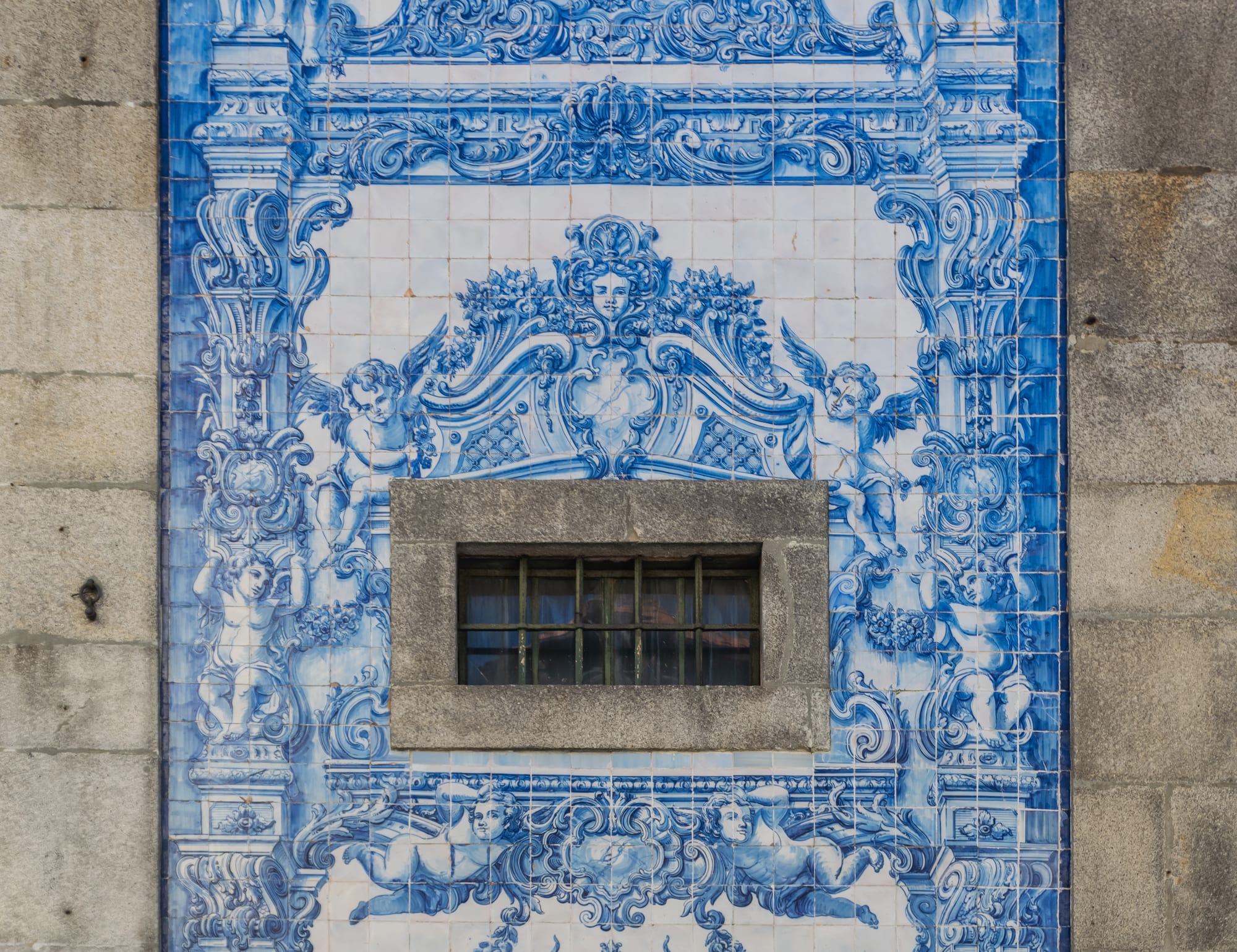 A wall of blue and white glazed tiles painted with baroque scrollwork, cherubs and a carved face, framing a small barred window set into the granite