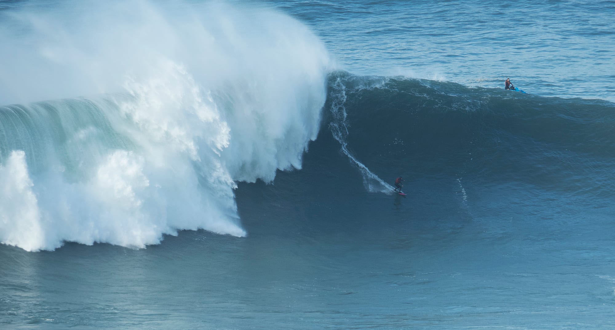 A tiny surfer riding a massive breaking wave, dwarfed by the wall of water