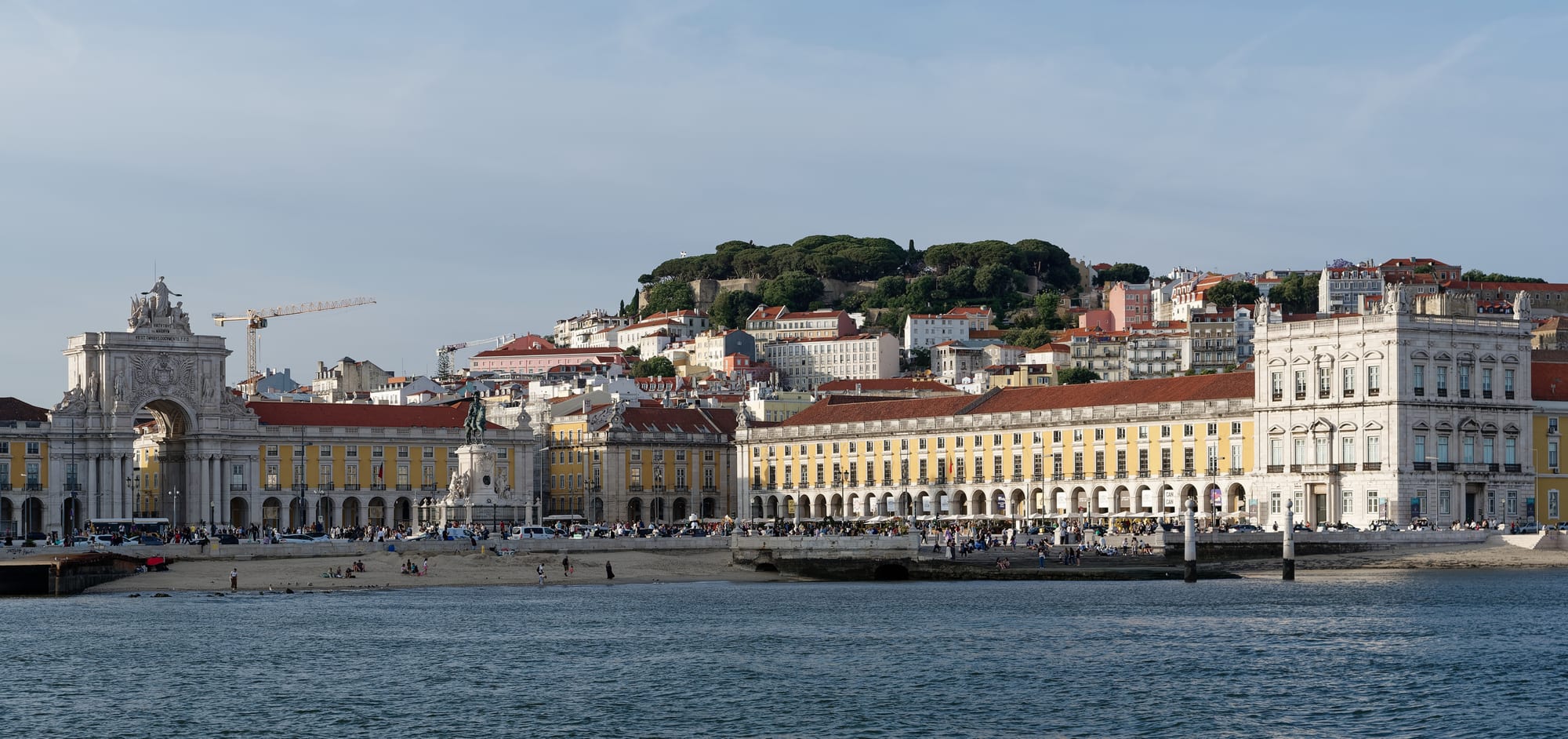 A grand riverfront square with a triumphal arch and colourful buildings, seen from the water