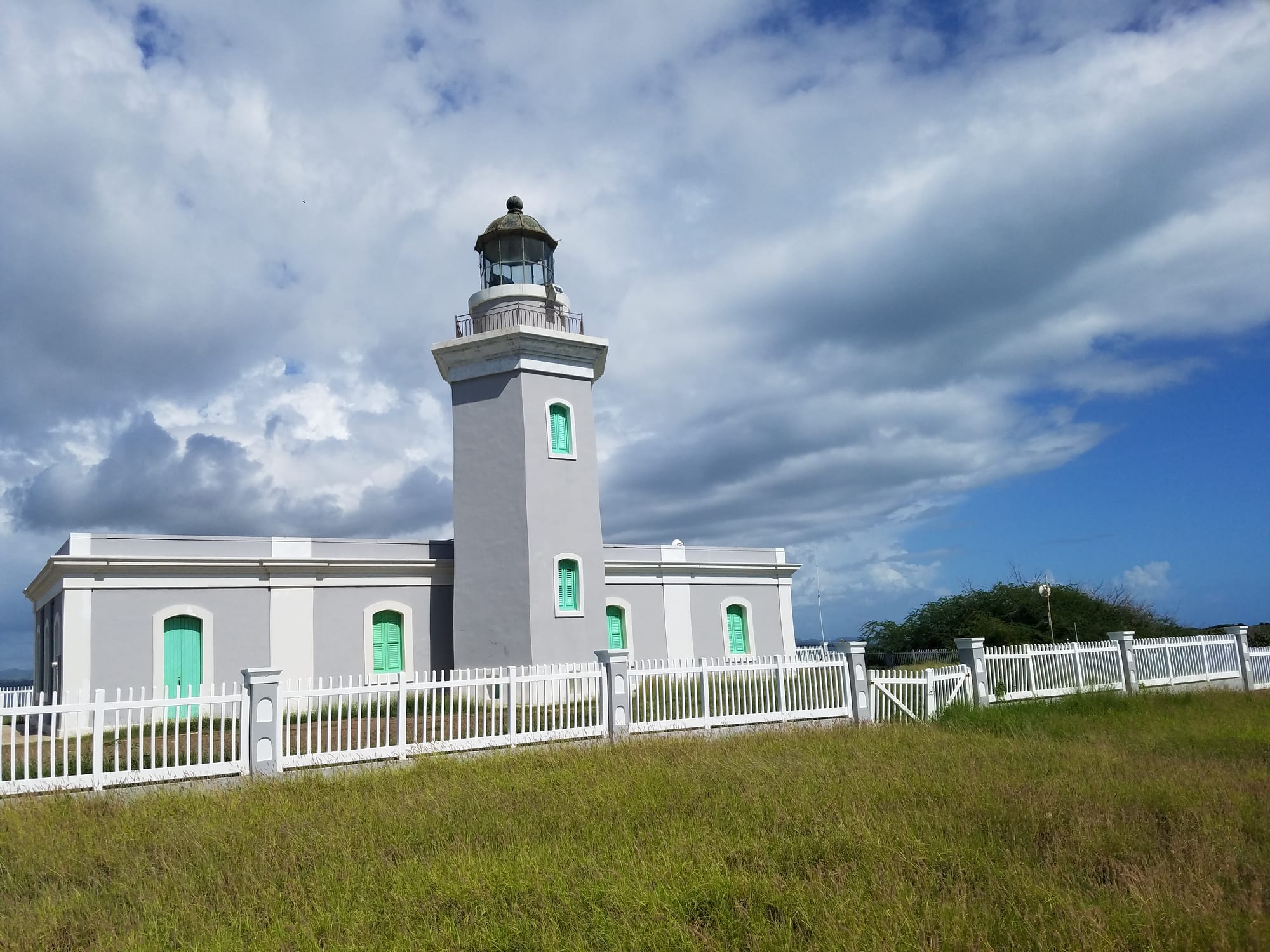A grey lighthouse with green-shuttered windows on a grassy headland under a partly cloudy sky