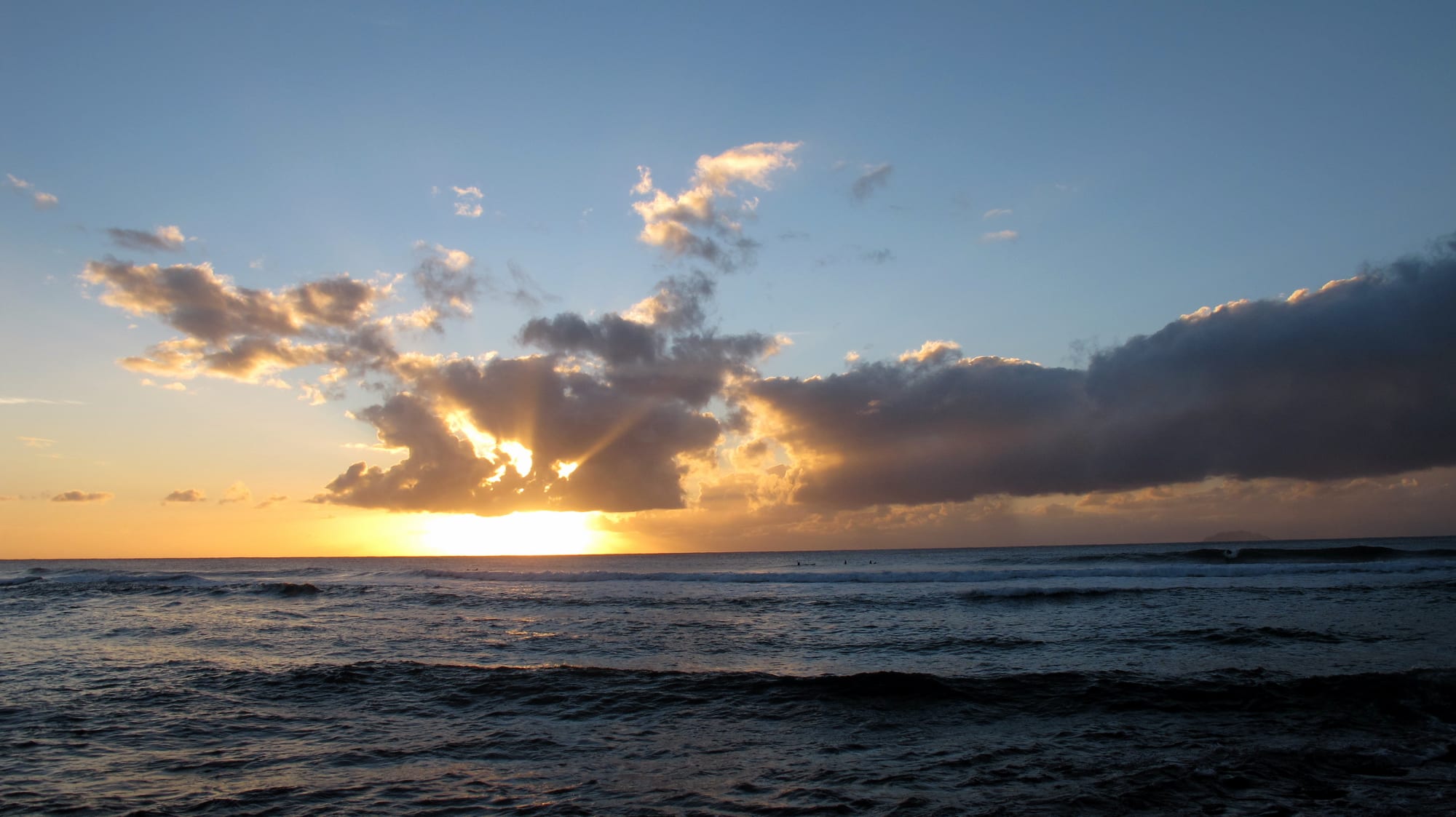A dramatic sunset with sun rays breaking through clouds over the ocean, distant surfers visible