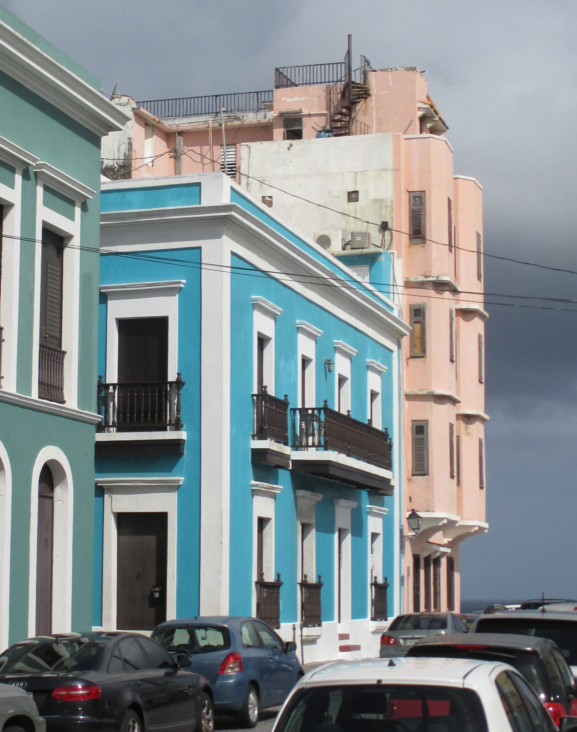 A blue Spanish colonial-style residential building with white trim and wrought-iron balconies, cars parked outside