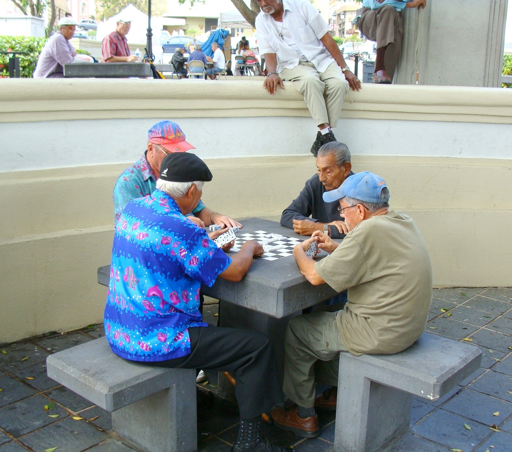 Four older men playing dominoes at a stone table in a plaza, with more people sitting and talking on a nearby wall