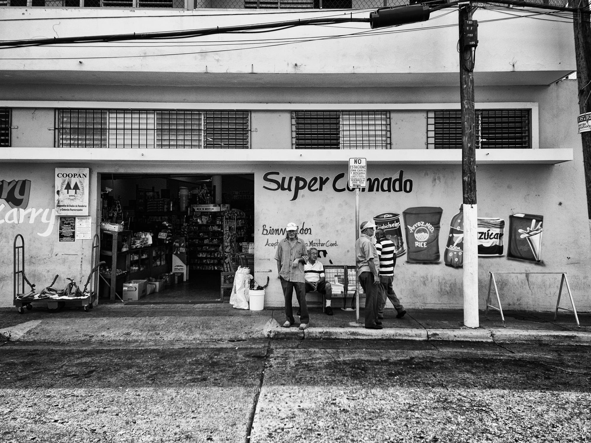 Men standing and sitting outside a corner grocery store called Super Colmado, painted signs for rice and sugar on the wall