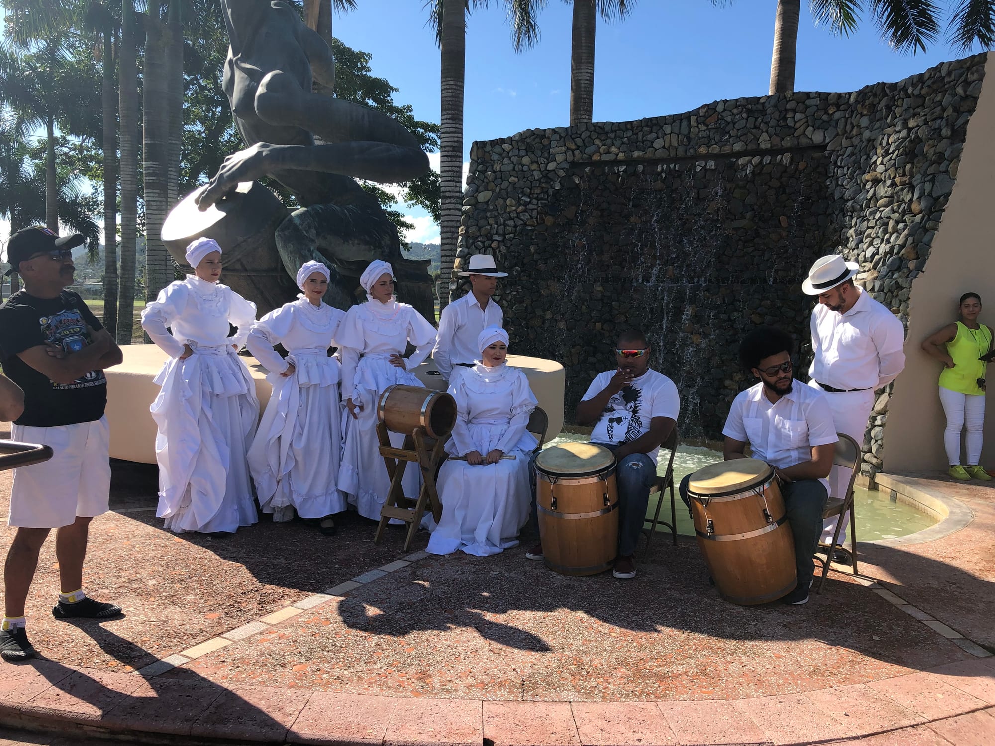 Four women in long white ruffled dresses and white headwraps stand beside three men seated at wooden barrel drums, with more musicians in white and straw hats, beside a large bronze sculpture and palm trees