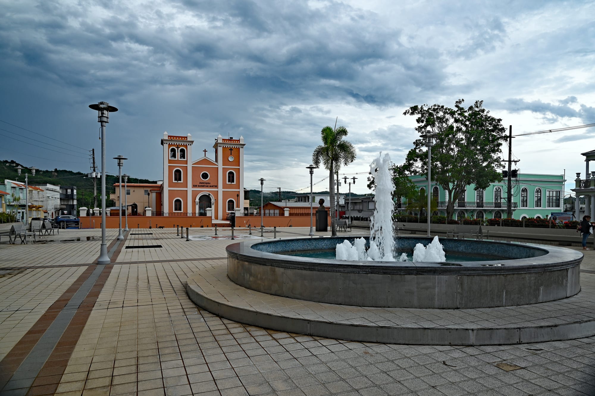 A wide tiled town square with a round fountain in the foreground, lamp posts and a palm tree, and a salmon-coloured church with two white-trimmed bell towers at the far end under a cloudy sky