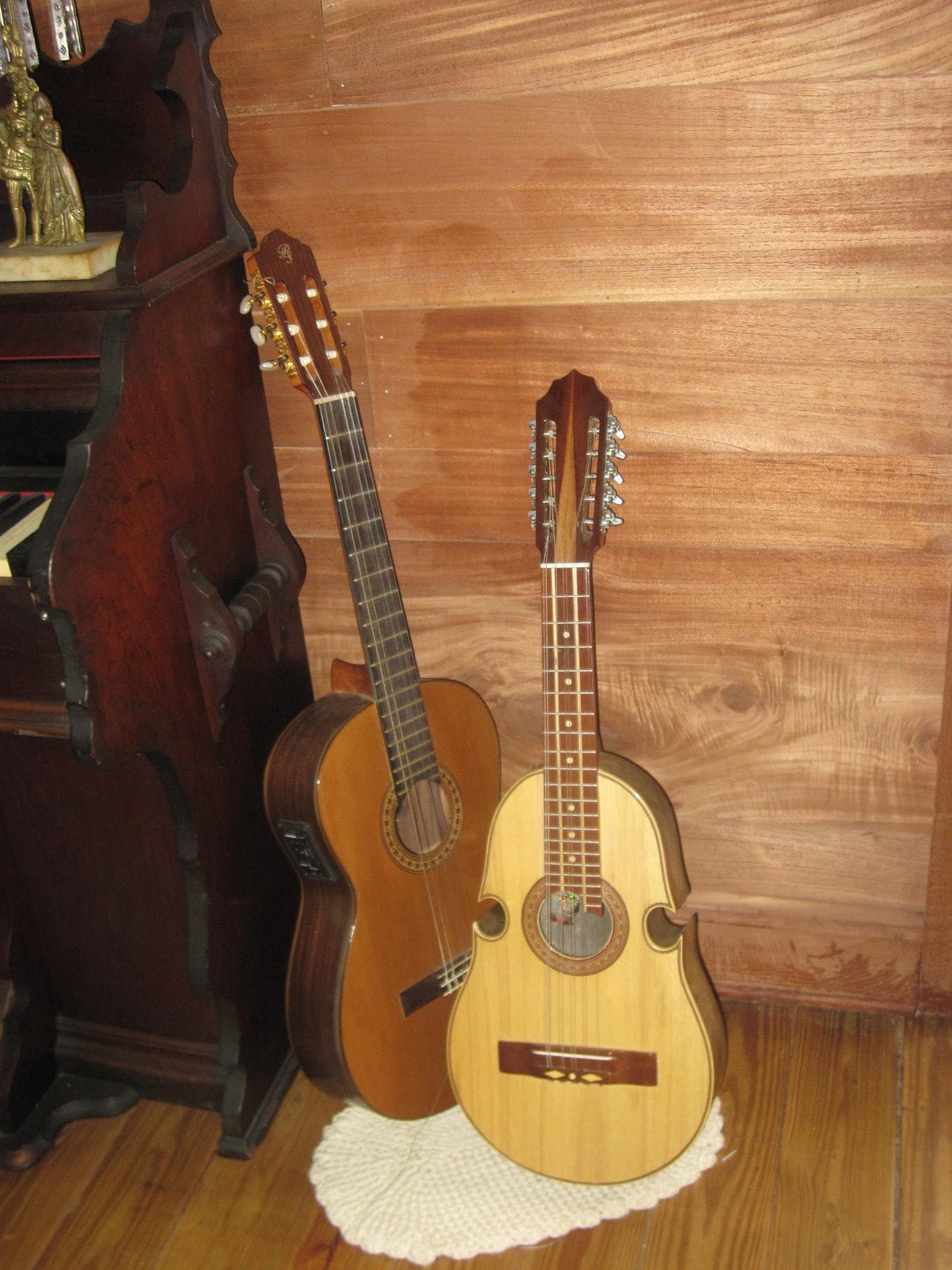 A Puerto Rican cuatro with a pale wooden body, a narrow waist and ten tuning pegs leaning against a classical guitar beside an old upright piano and a wood-panelled wall
