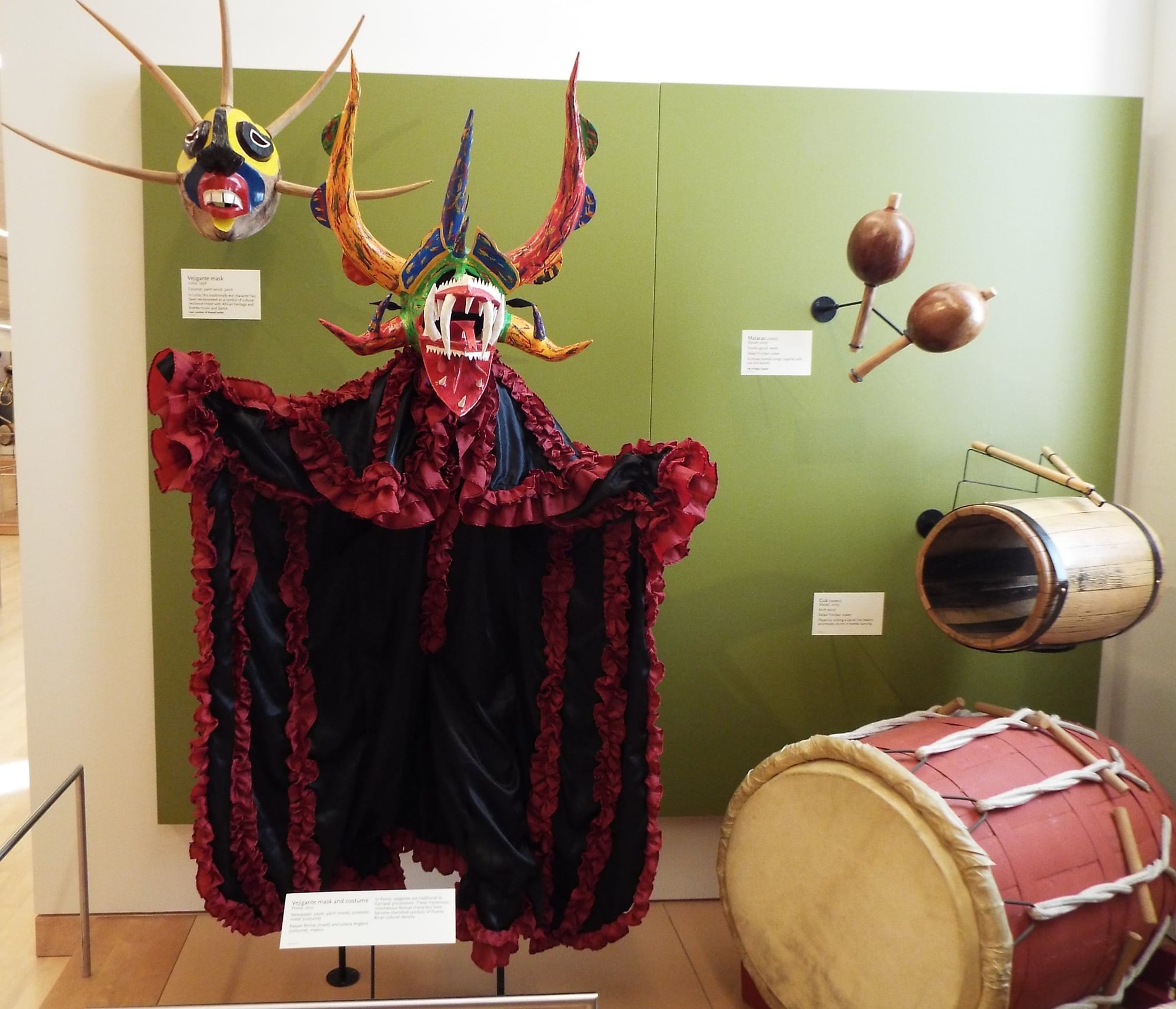 A museum display of a large horned vejigante mask in red, yellow and green with bared teeth above a black and red ruffled costume, with a smaller horned mask, a pair of maracas and two barrel drums beside it