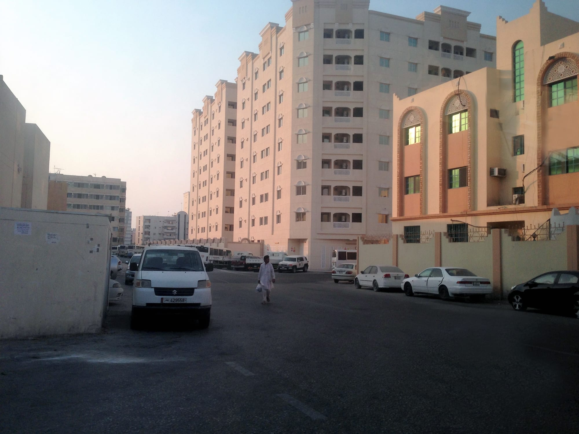 A street of cream and pink apartment buildings in a Doha residential district, with parked cars and a person in white robes walking