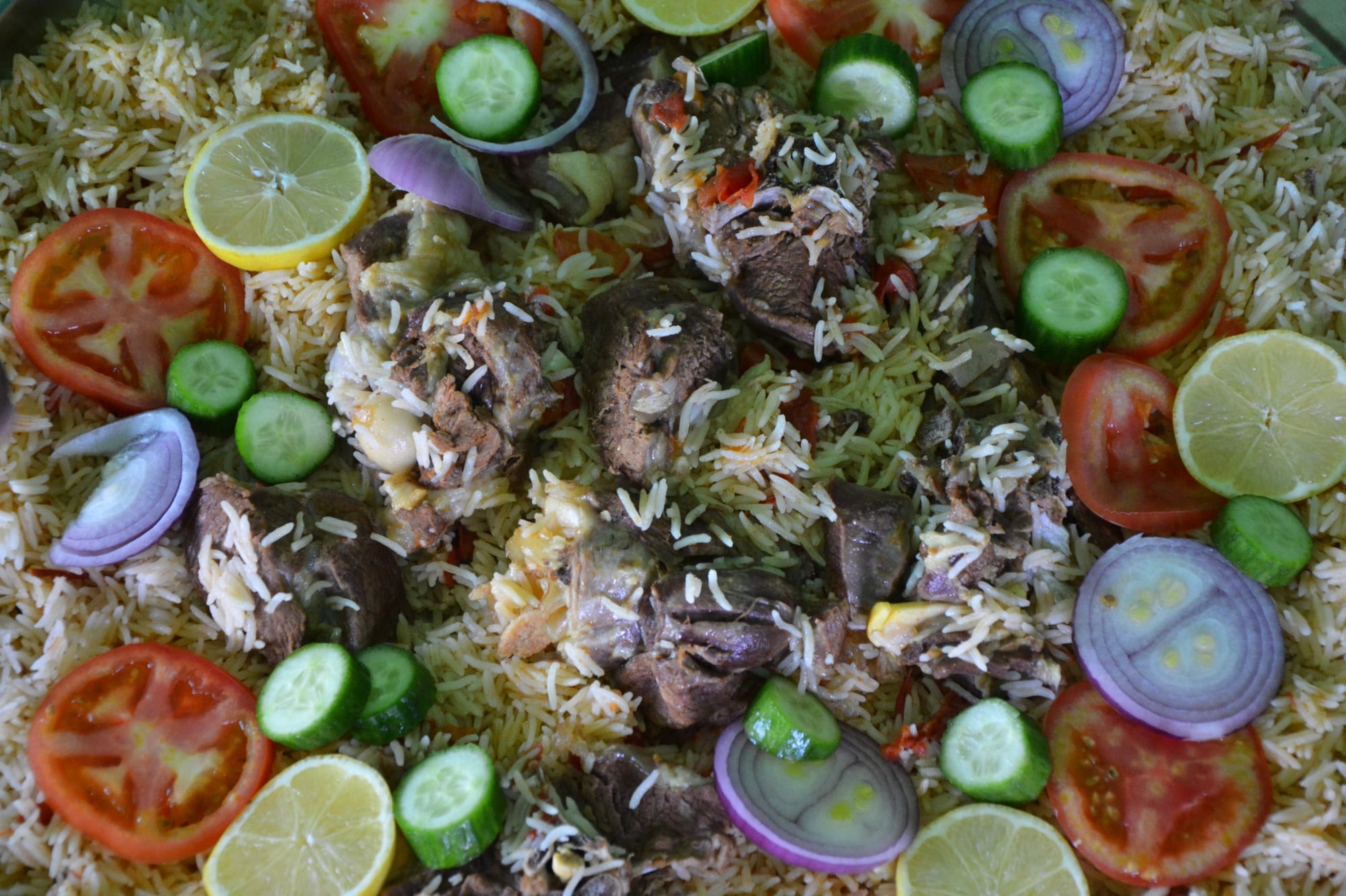 A close view of spiced rice topped with chunks of meat on the bone and garnished with slices of tomato, cucumber, lemon and red onion