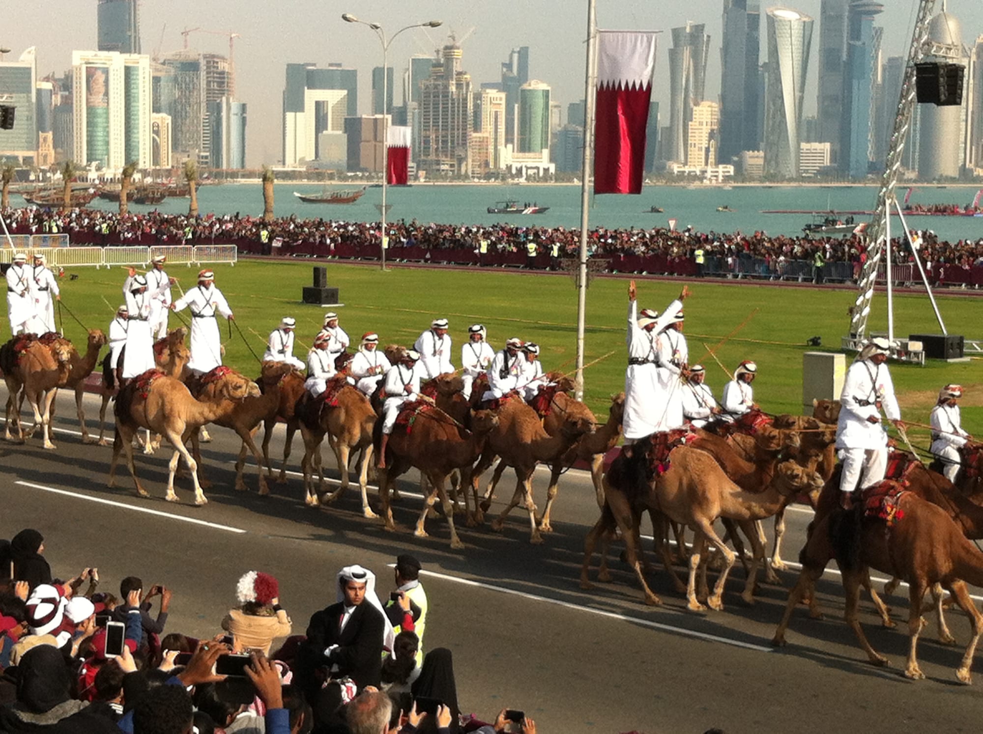 Rows of riders in white robes and red and white headdresses lead camels along a road beside a grass field, watched by large crowds, with maroon and white flags and a skyline of towers across the bay behind