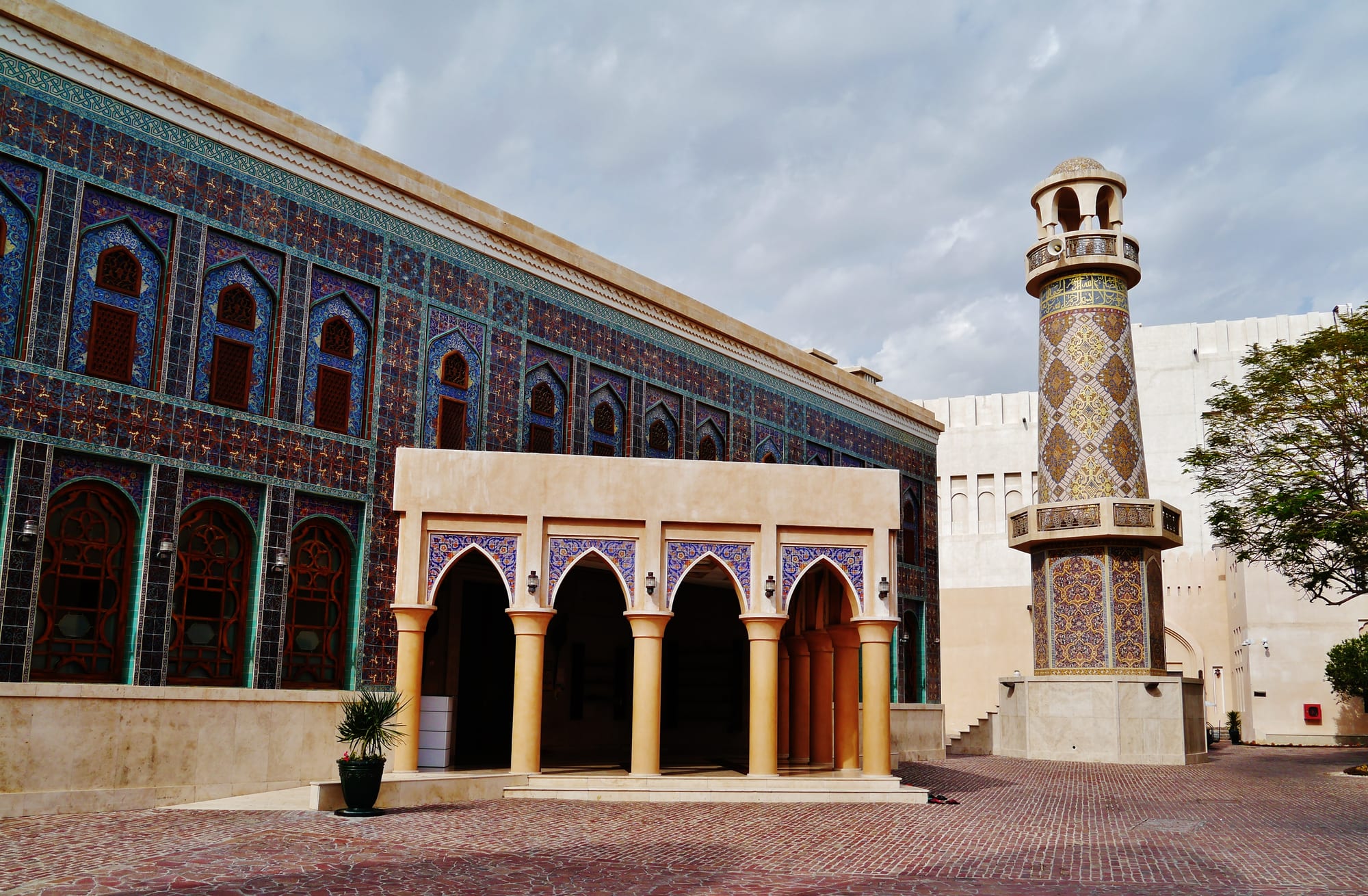 A mosque with walls covered in blue, purple and gold tiles in arched patterns, a pale arcaded porch at the front and a tall tiled minaret to the right, under a cloudy sky