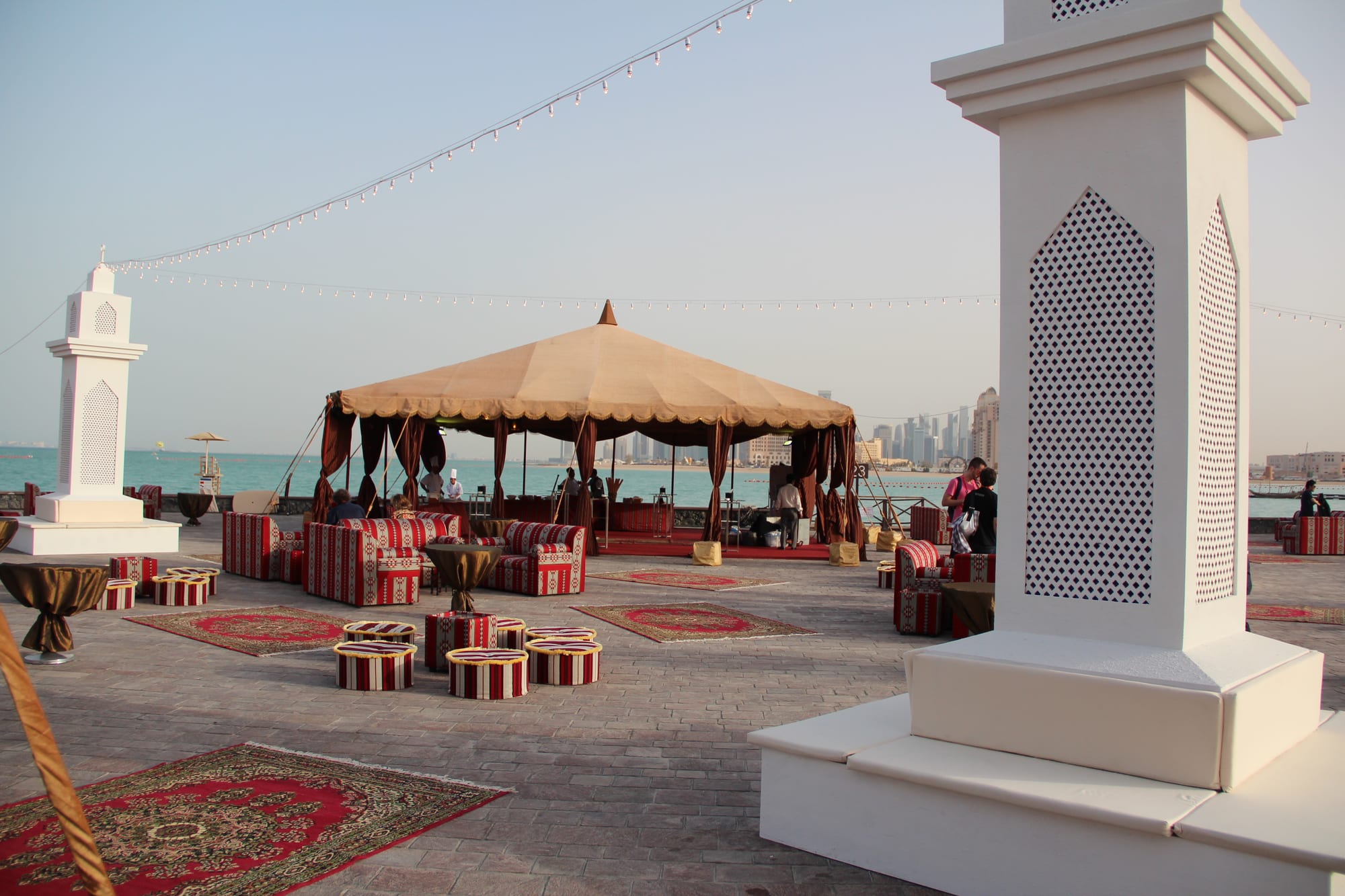 Low sofas and cushioned stools upholstered in red and cream stripes arranged on patterned rugs around a large beige canopy on a waterfront promenade, with the sea and a city skyline behind