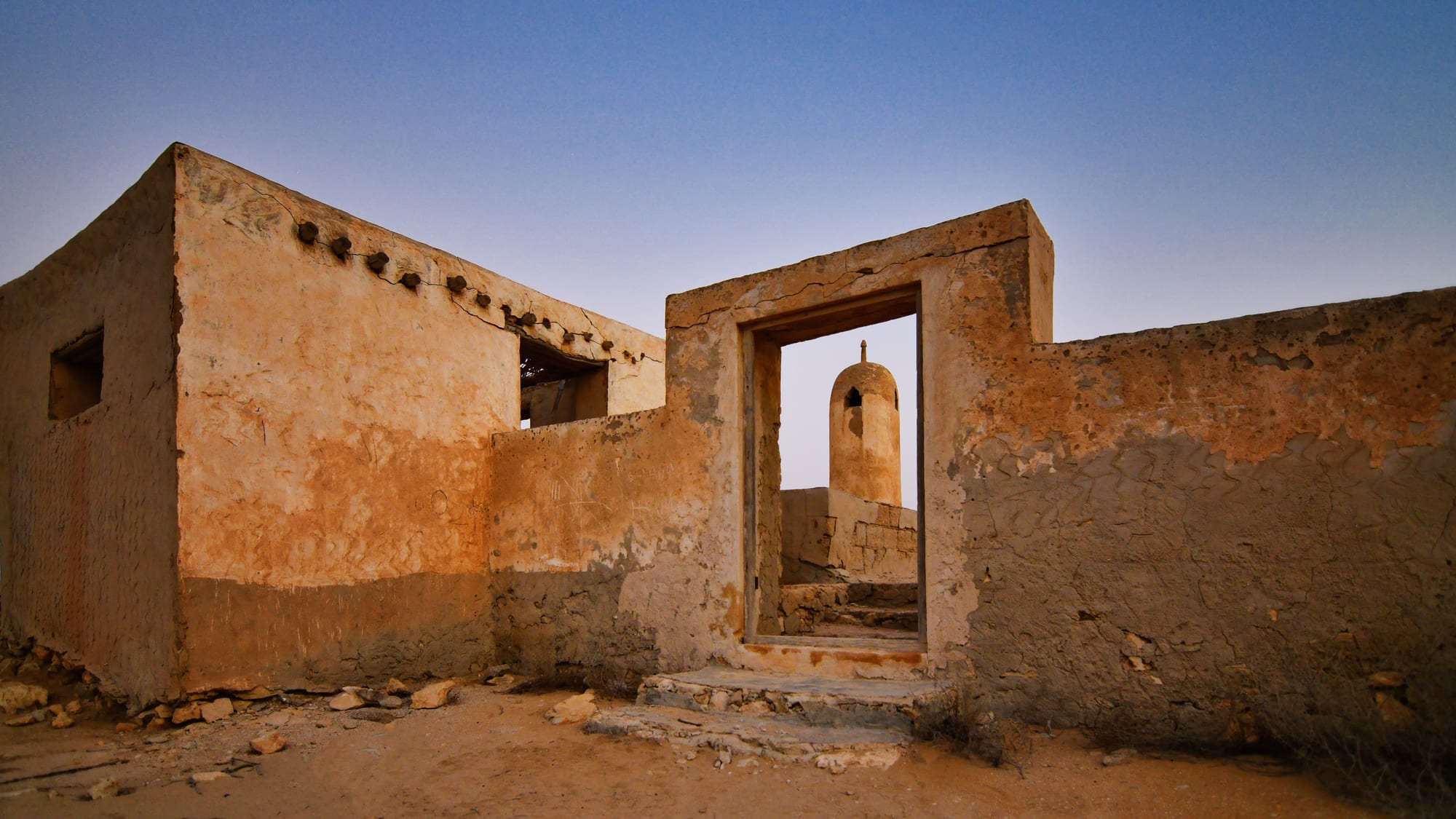 Roofless mud-plastered stone houses on sandy ground at dusk, with a small rounded minaret framed through an empty doorway