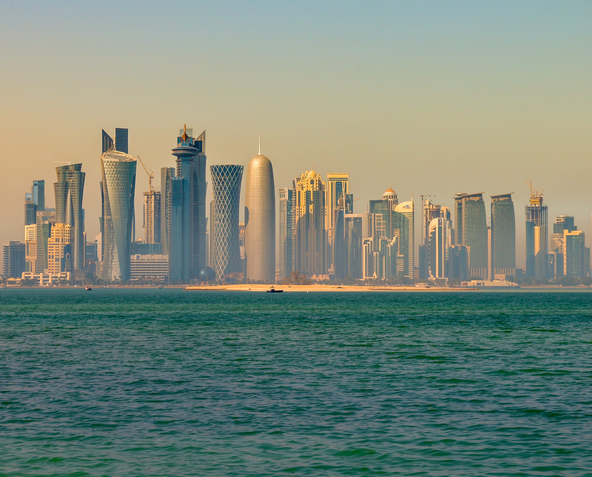 A row of glass skyscrapers of varied shapes rising along the waterfront of Doha at dawn