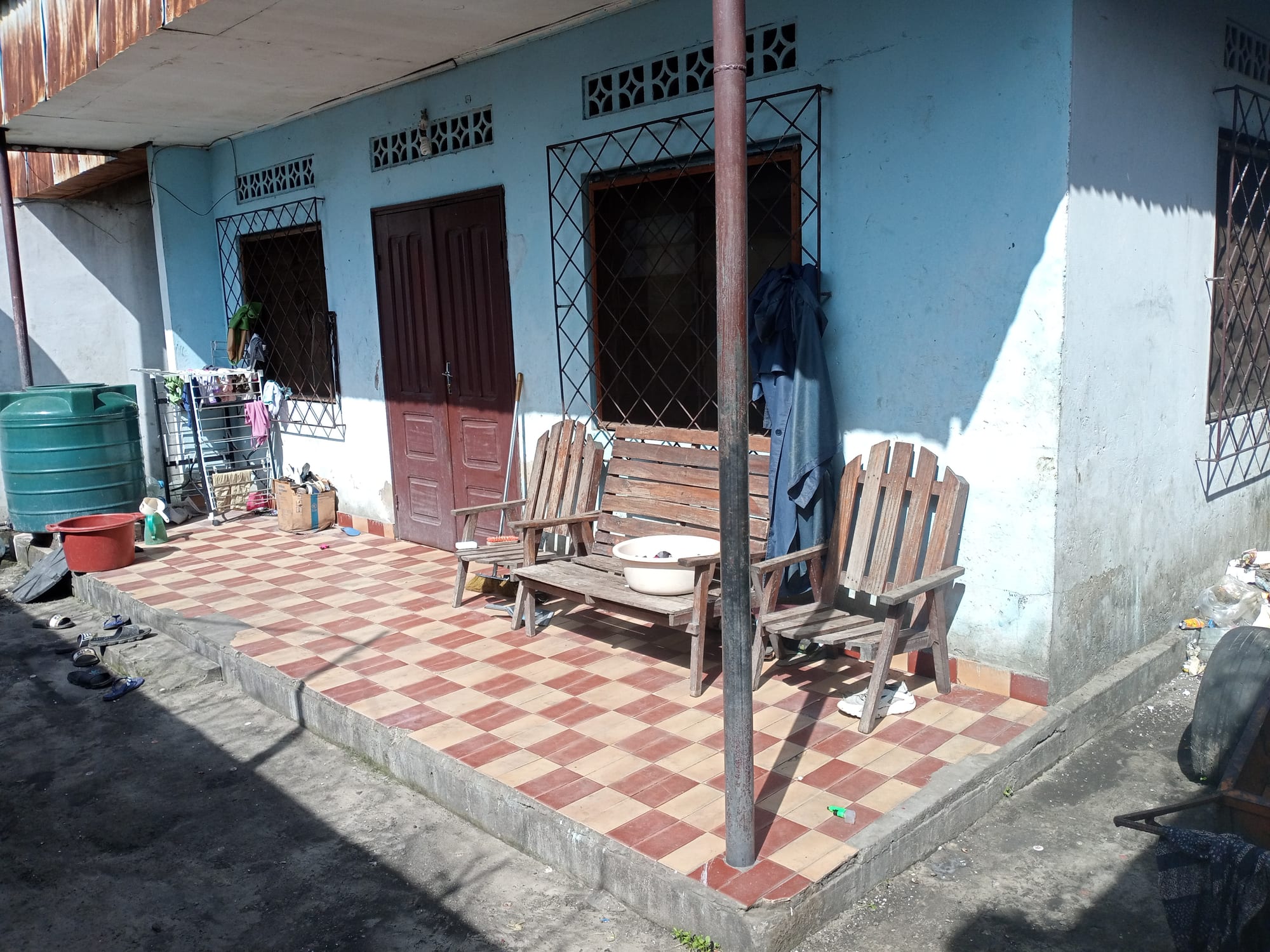 The tiled front porch of a light blue house, with wooden chairs, a bench, laundry rack and water containers