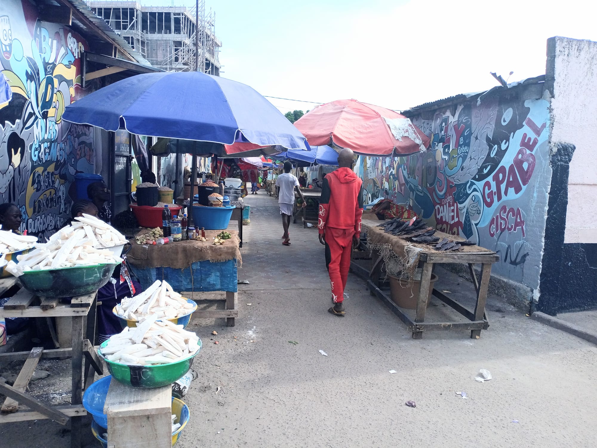 A narrow market alley in Brazzaville lined with colourful umbrellas, painted murals and stalls selling cassava and fish