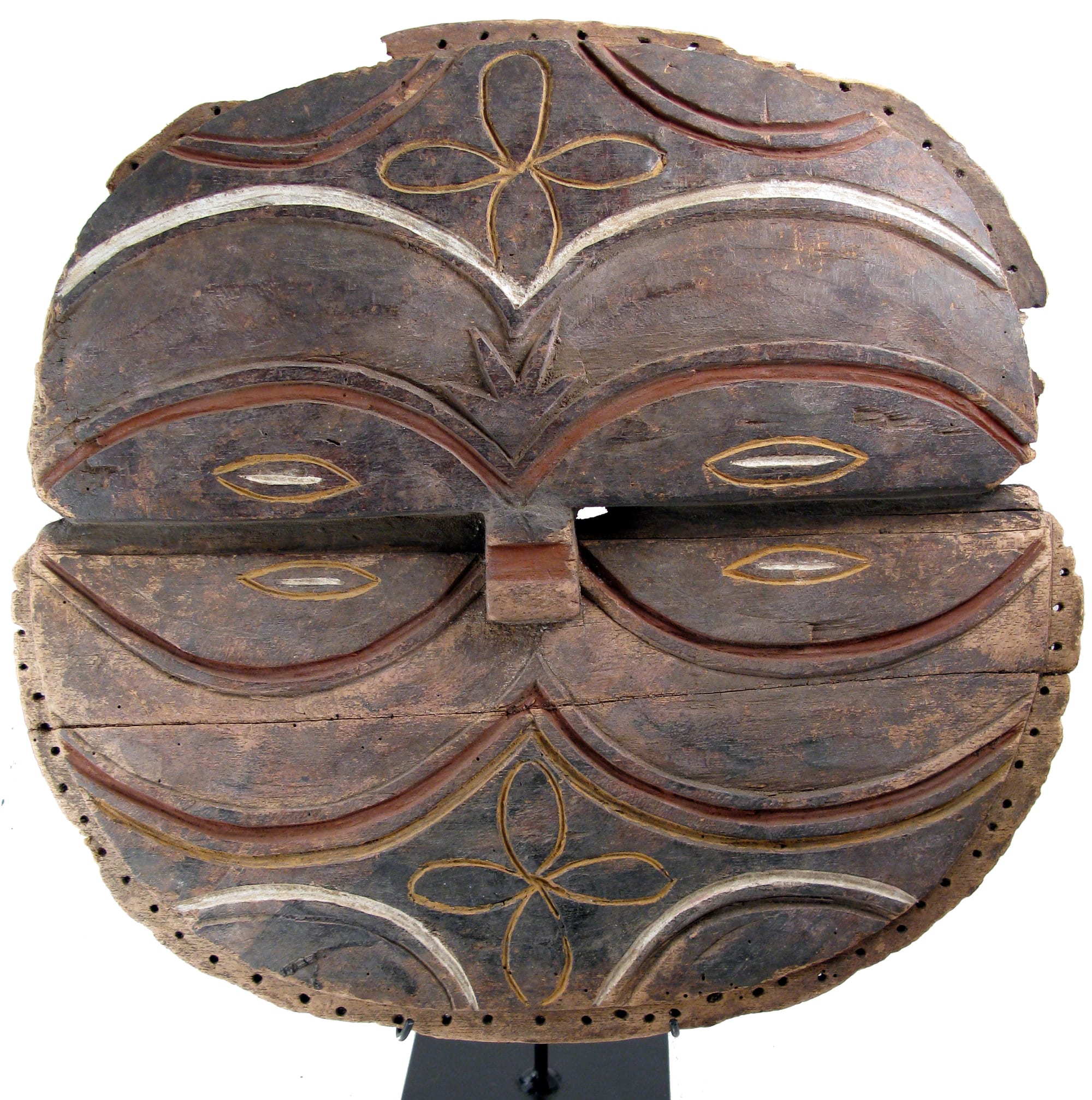 A round, flat wooden mask split by a horizontal groove across the middle, carved in symmetrical arcs outlined in red, white and yellow, with narrow almond-shaped eye slits, a small projecting nose and a ring of small holes around the rim