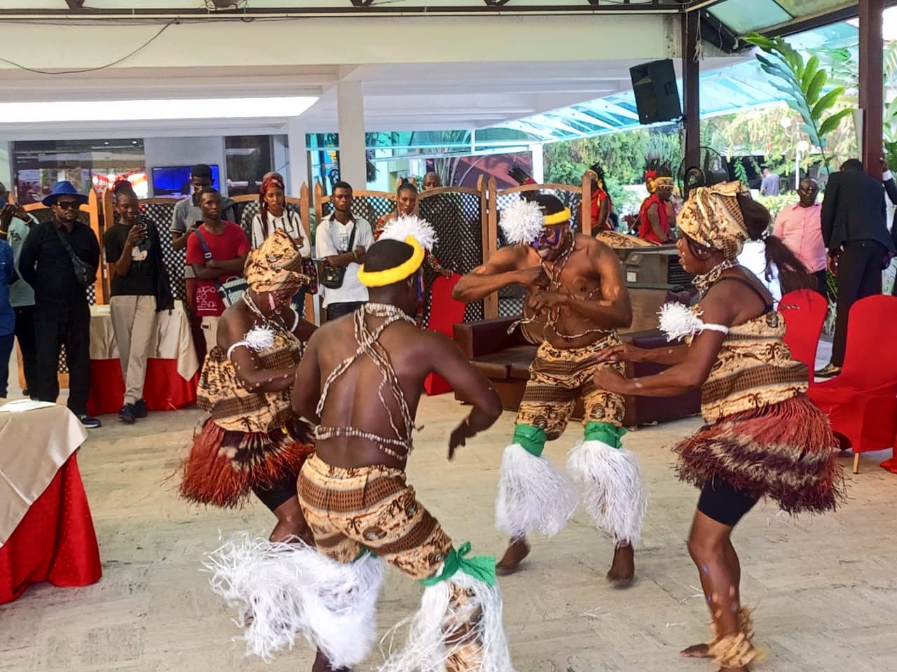 Four dancers in patterned costumes with raffia skirts, white fibre ruffs on their legs and yellow headbands dance energetically on a tiled floor as a crowd watches under a canopy