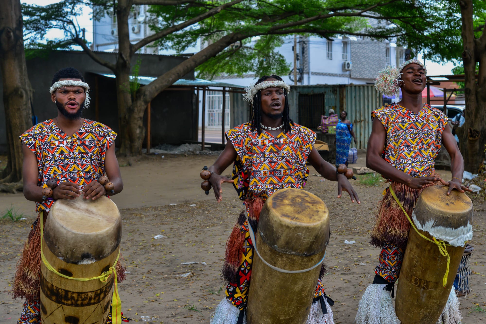 Three young men in matching sleeveless tunics printed with orange, red and black patterns, raffia skirts and white fibre headbands sing as they play tall wooden drums slung from their shoulders, under shady trees