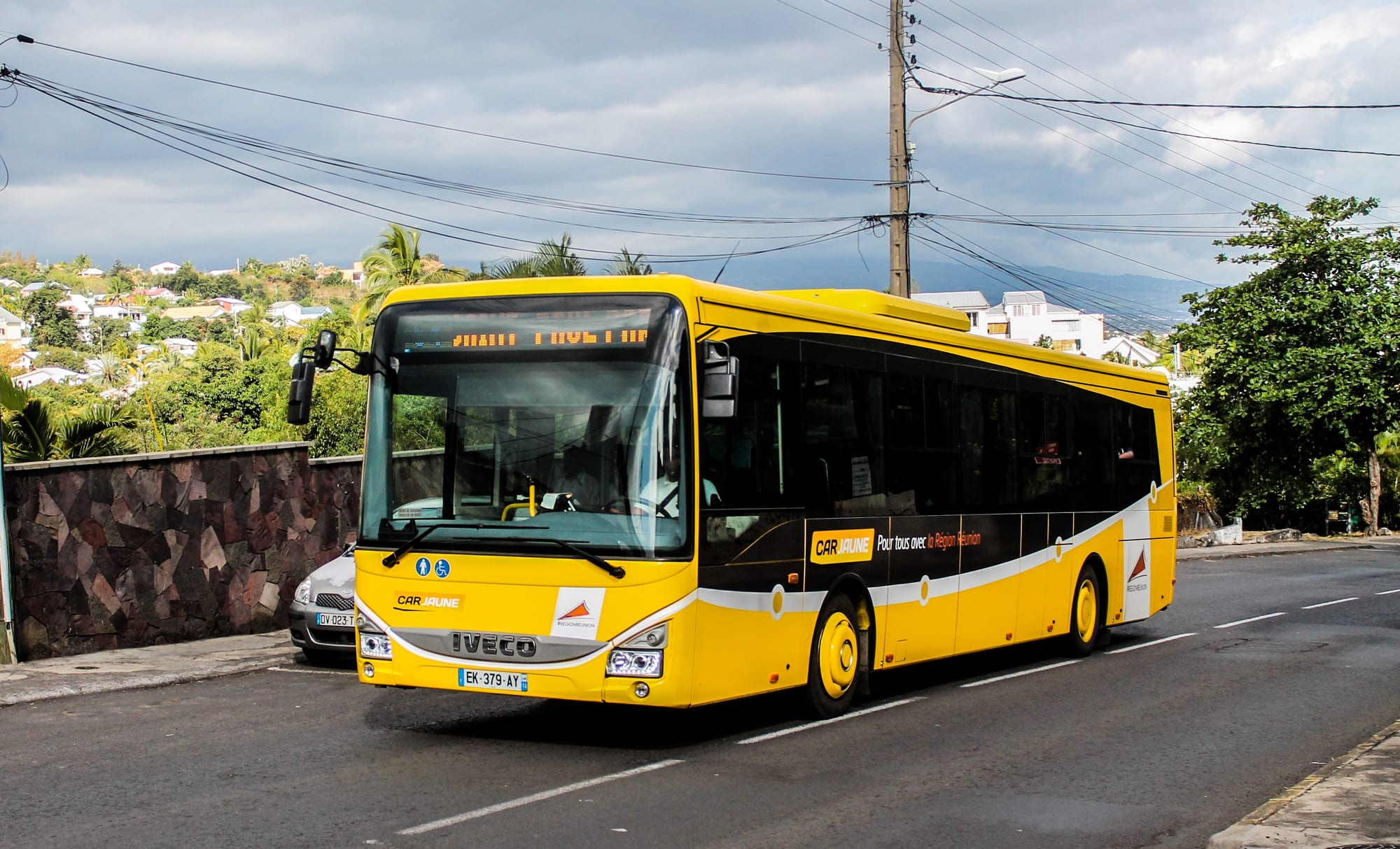 A yellow Car Jaune bus driving along a coastal road with hillside houses in the background