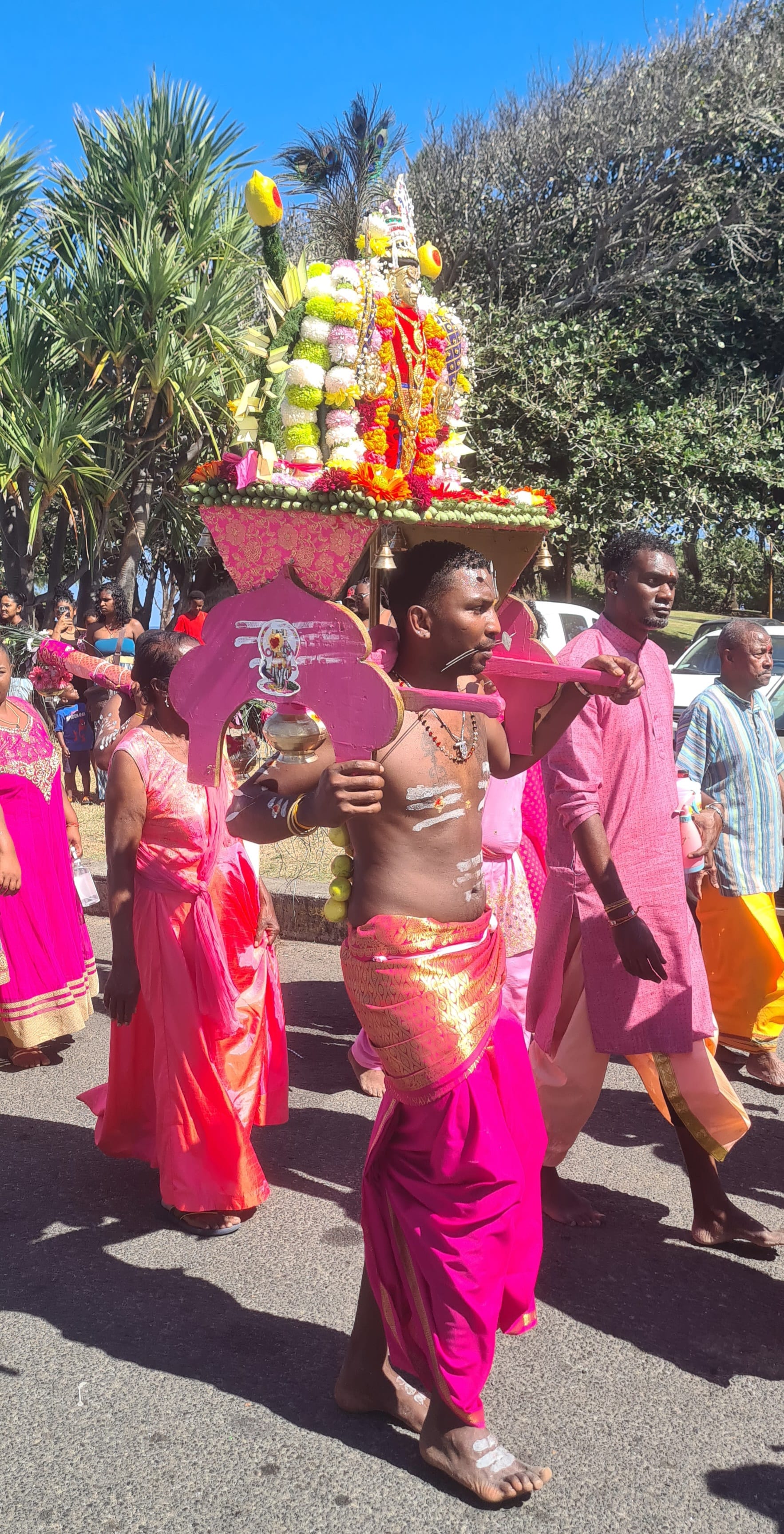A barefoot man in a pink and gold dhoti carries a pink wooden kavadi topped with flowers and a small statue, walking in a street procession with women in bright saris