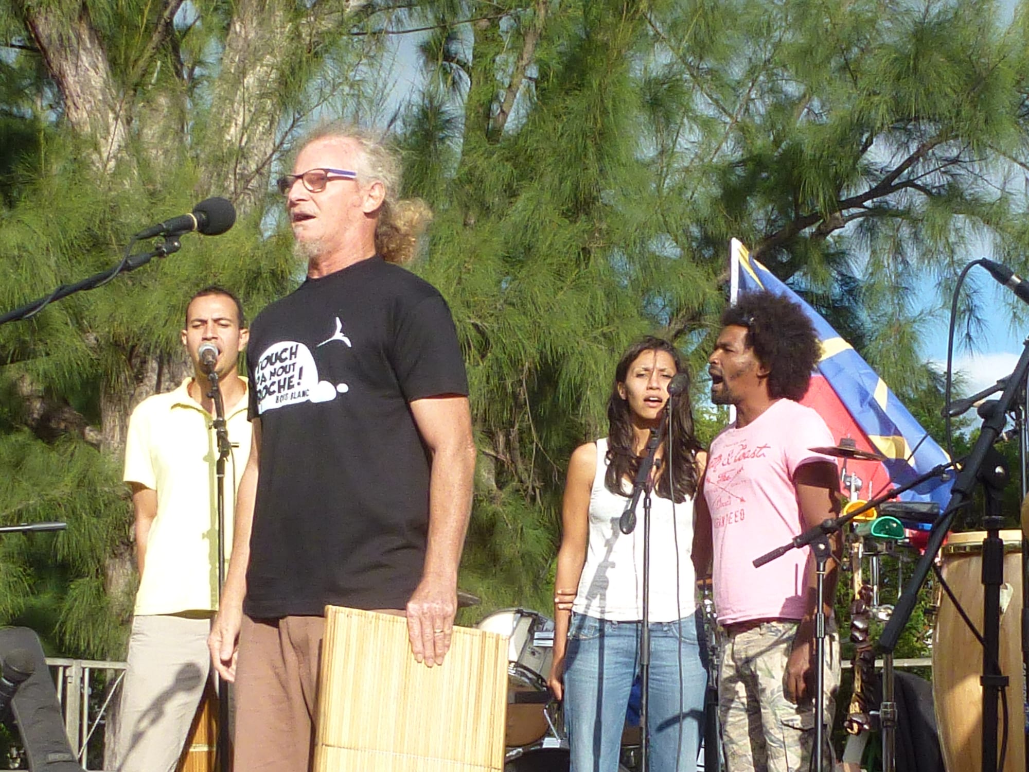 A grey-haired singer in glasses and a black T-shirt sings at a microphone holding a flat rectangular rattle, with three other singers and drums behind him under tall trees
