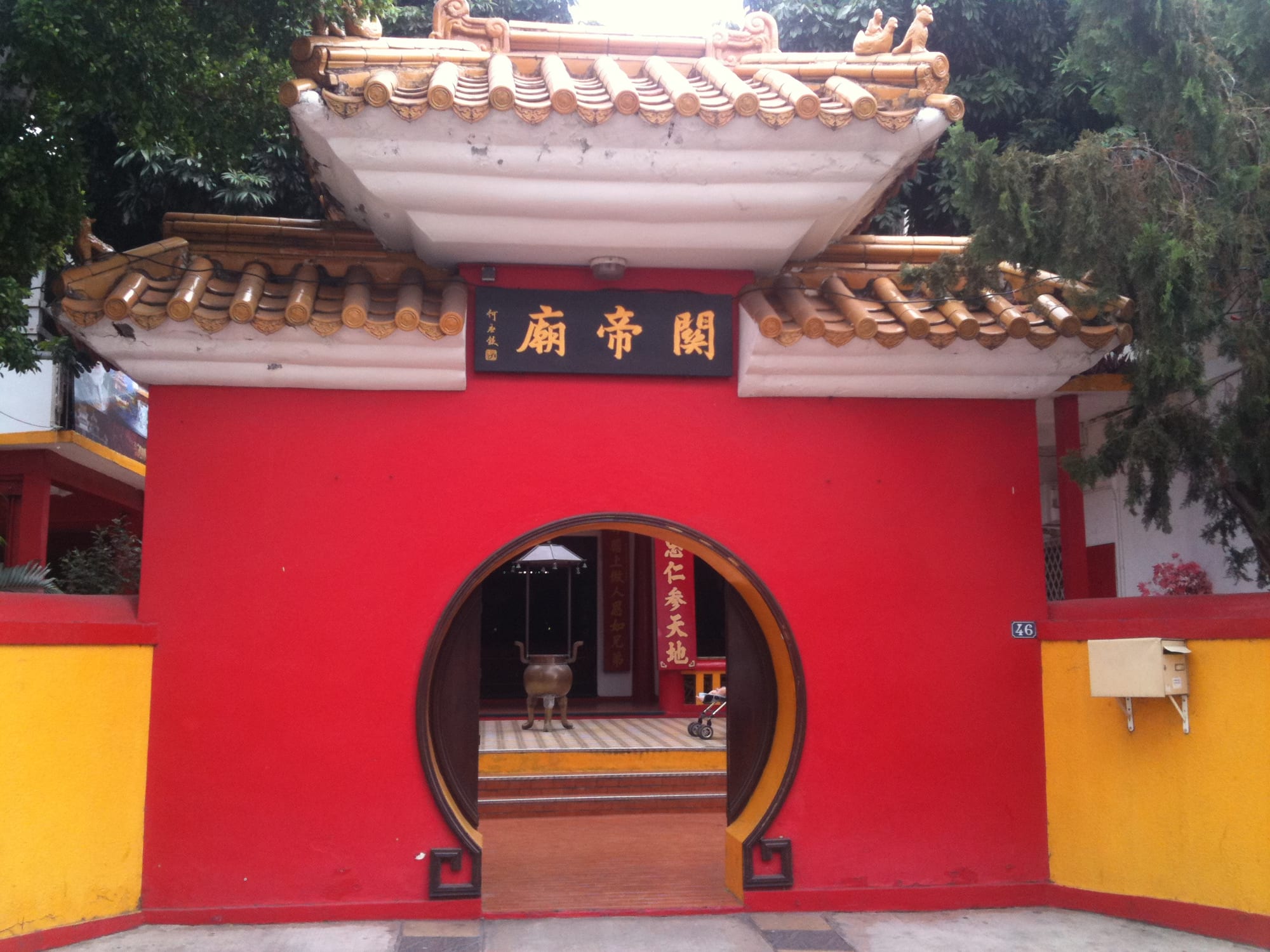 A red wall with a round moon-gate doorway beneath curved golden-tiled roofs and a black plaque bearing three Chinese characters, with a bronze incense burner visible through the opening