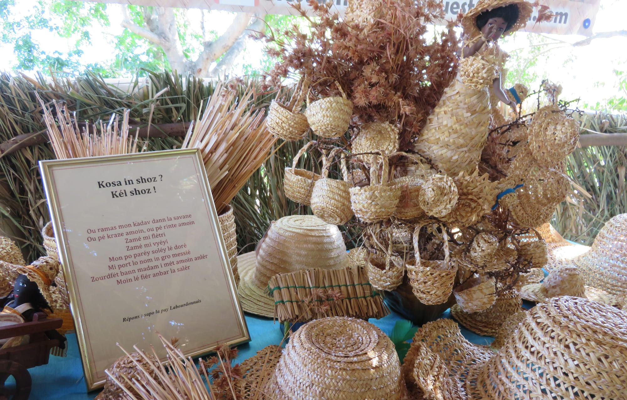 A table display of woven straw baskets, hats, bags and a doll in a woven dress, with bundles of dried grass stems and a framed Creole poem at the front