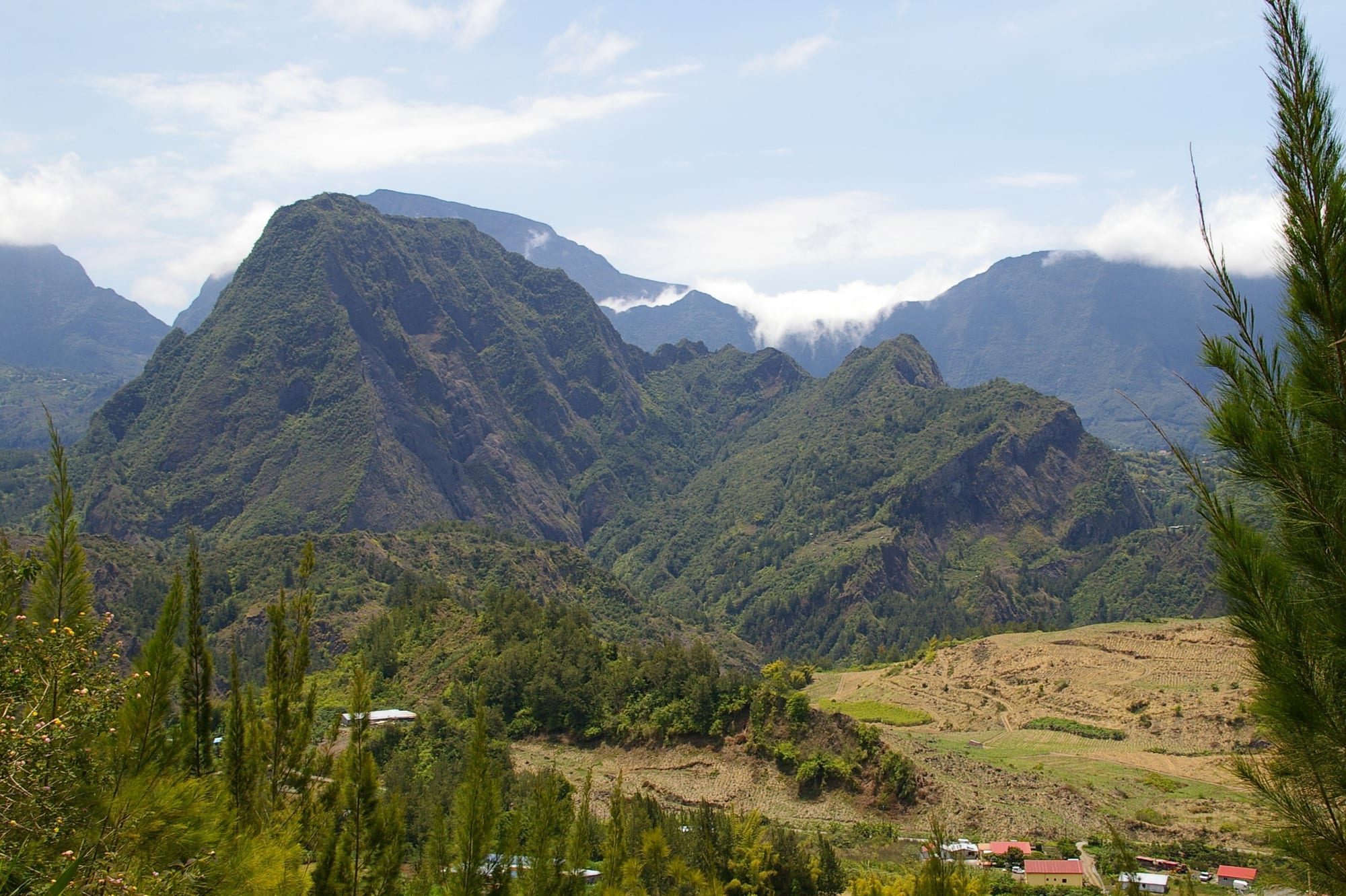 A steep, forest-covered peak rising from a green valley with a few small houses below and cloud-capped mountain walls behind
