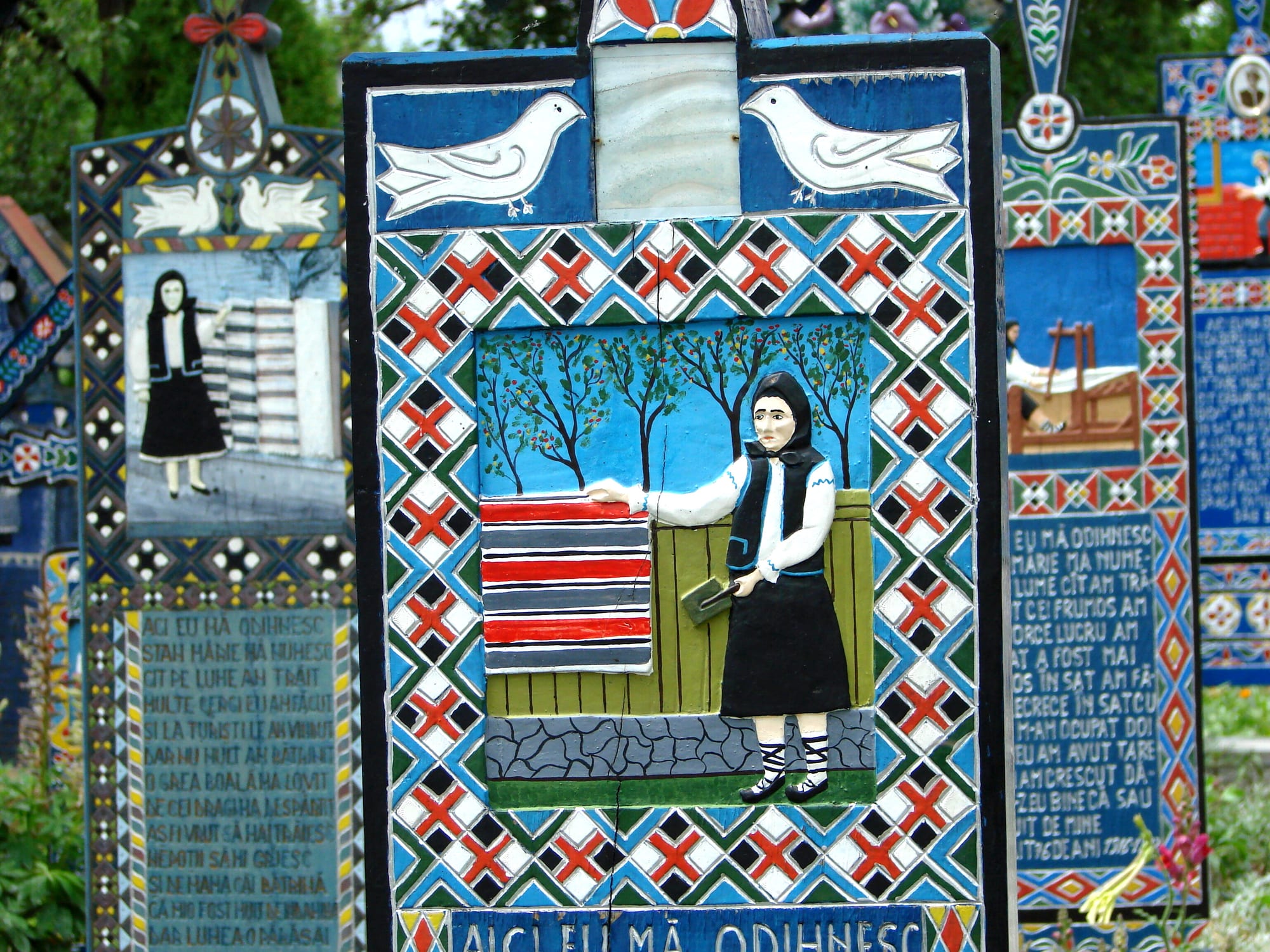 A carved wooden grave cross painted in bright blue with red and black patterns, white doves at the top and a relief of a woman beside a striped woven cloth, with other painted crosses behind