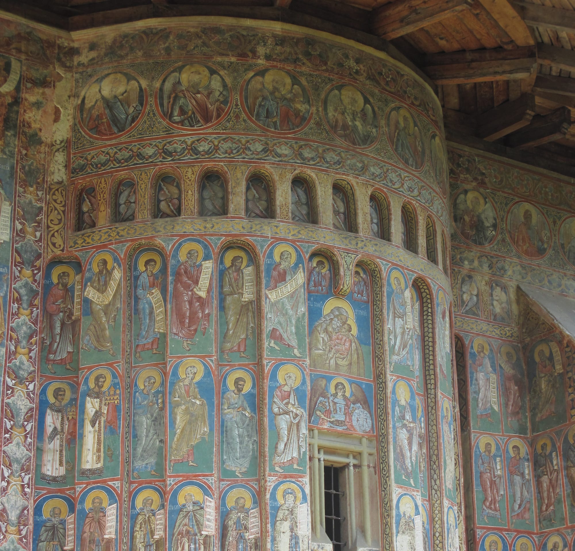 The curved outer wall of a church covered from top to bottom in painted rows of haloed saints and angels in red, blue and gold, beneath a wooden roof