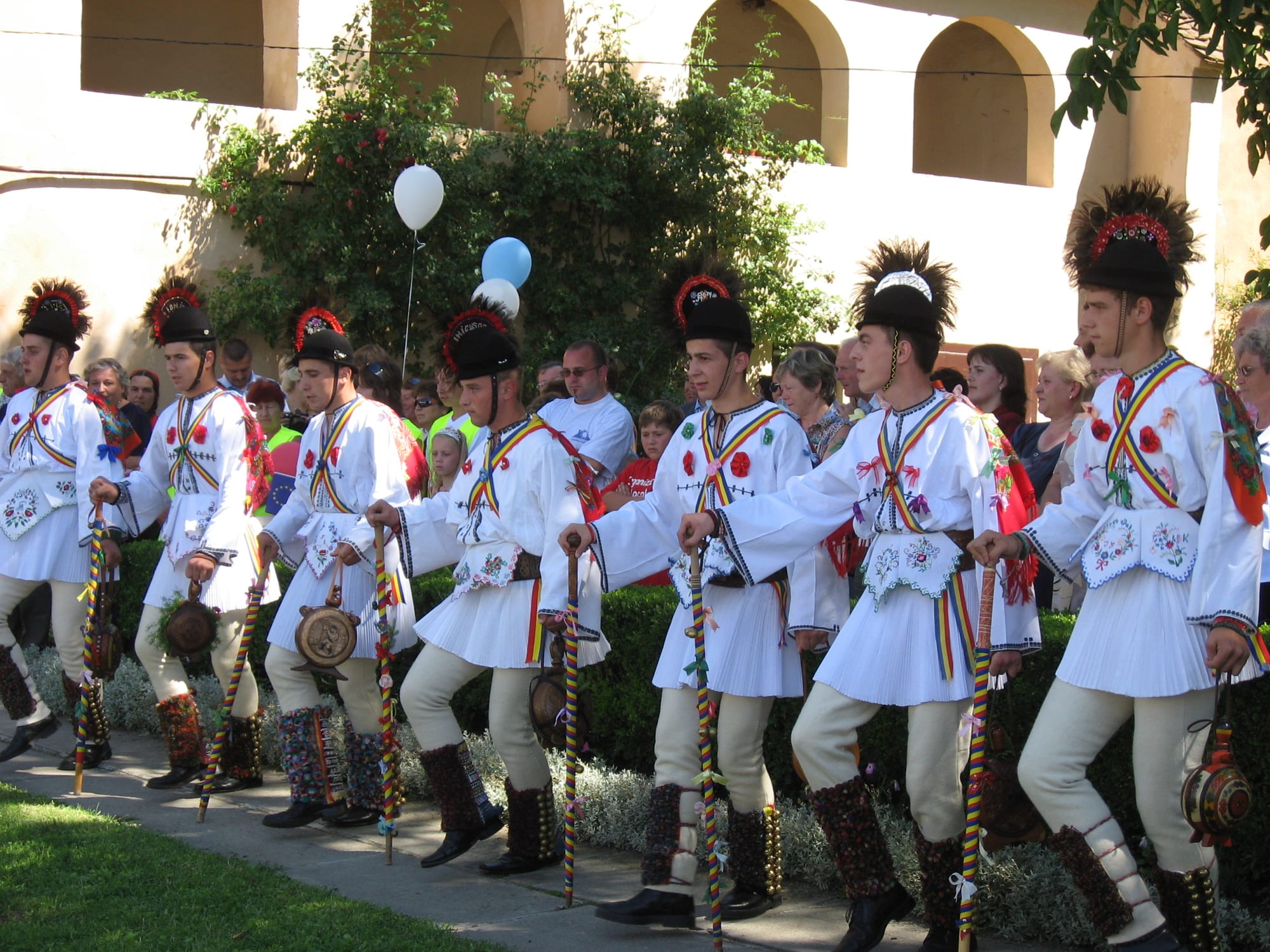 A line of young men in white pleated costumes, black hats with red plumes and striped sashes stand holding decorated sticks in front of a crowd