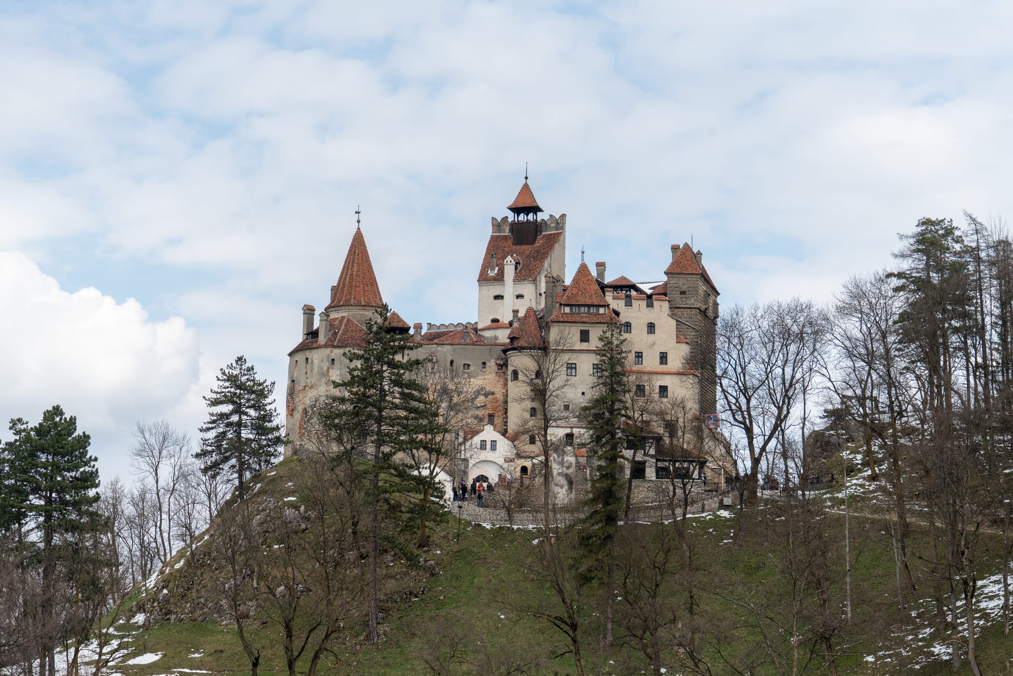 A tall white medieval castle with red roofs perched on a rocky outcrop above forest in Transylvania