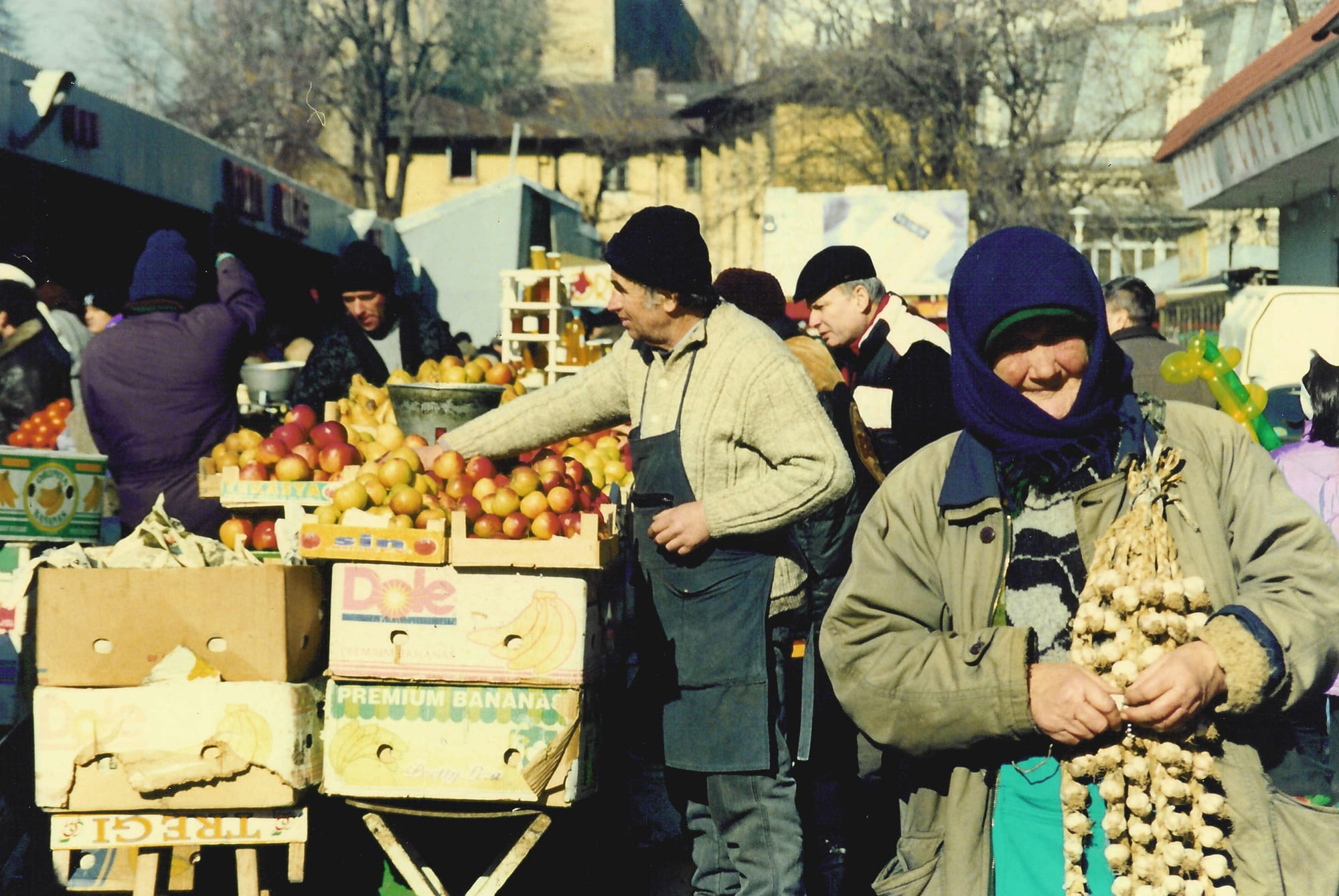 A fruit vendor arranging apples in crates and a woman holding strings of garlic at an open-air produce market