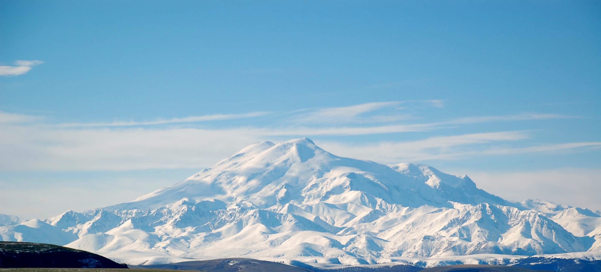 A massive twin-peaked snow-covered volcano rising above lower mountains