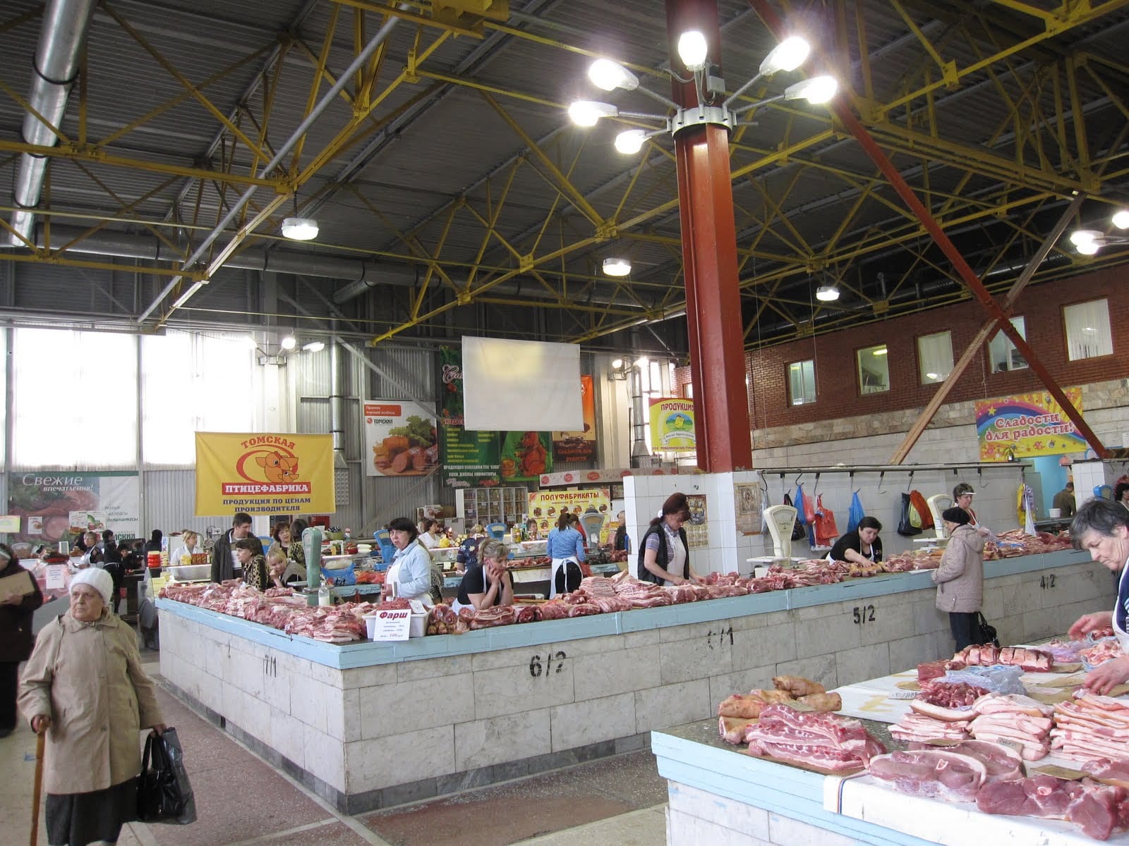 Shoppers and vendors at butcher counters inside a large covered market hall in Tomsk