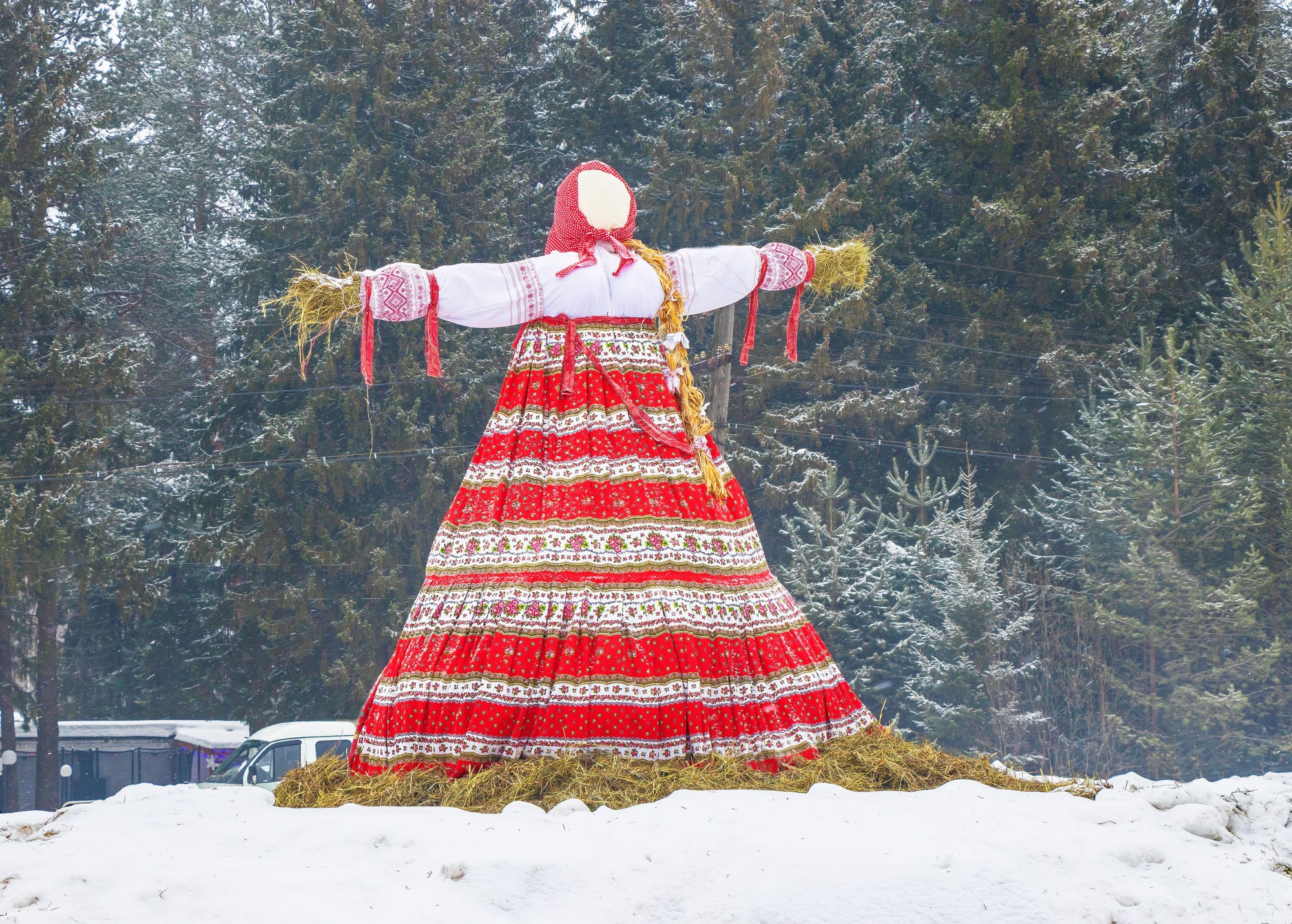 A tall straw effigy in a white embroidered blouse, red headscarf and long red and white patterned skirt stands on a bed of straw in the snow, with fir trees behind