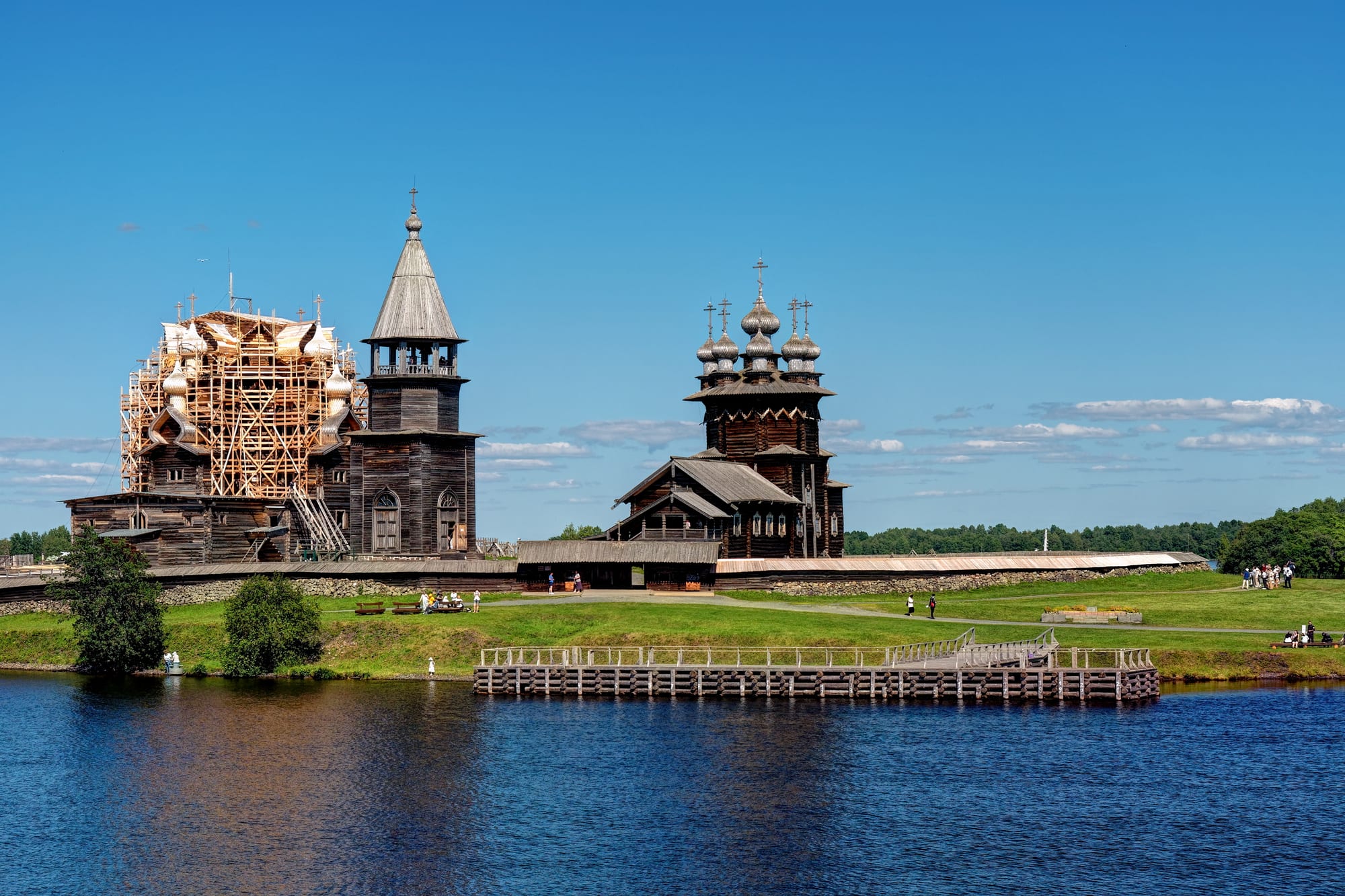 Two dark wooden churches with clusters of onion domes and a tall wooden bell tower stand on a green lakeside under a blue sky, one church wrapped in restoration scaffolding