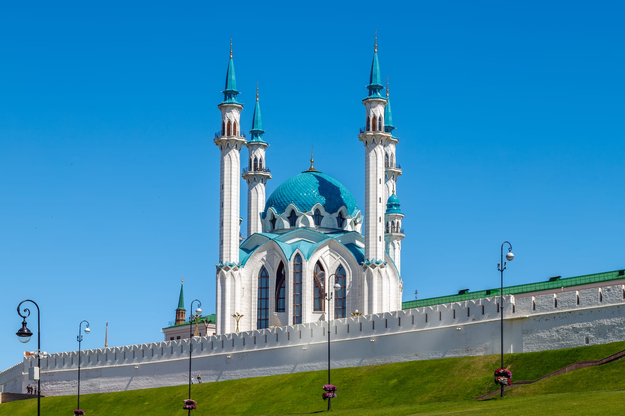 A white mosque with a turquoise dome and four tall white and turquoise minarets rising above a long white crenellated wall and a grassy slope