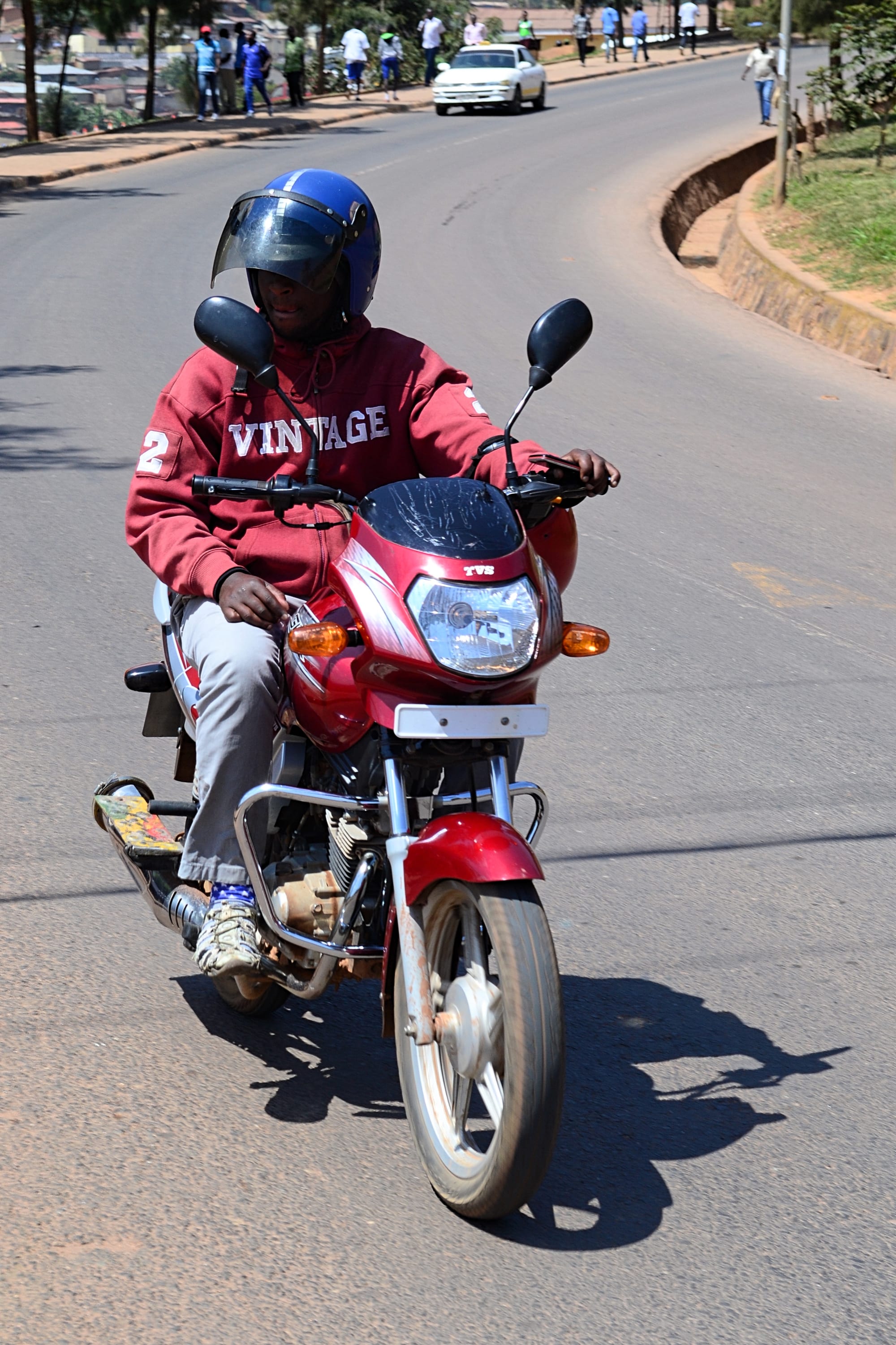 A helmeted motorcycle taxi rider carrying a passenger along a curving road in Kigali