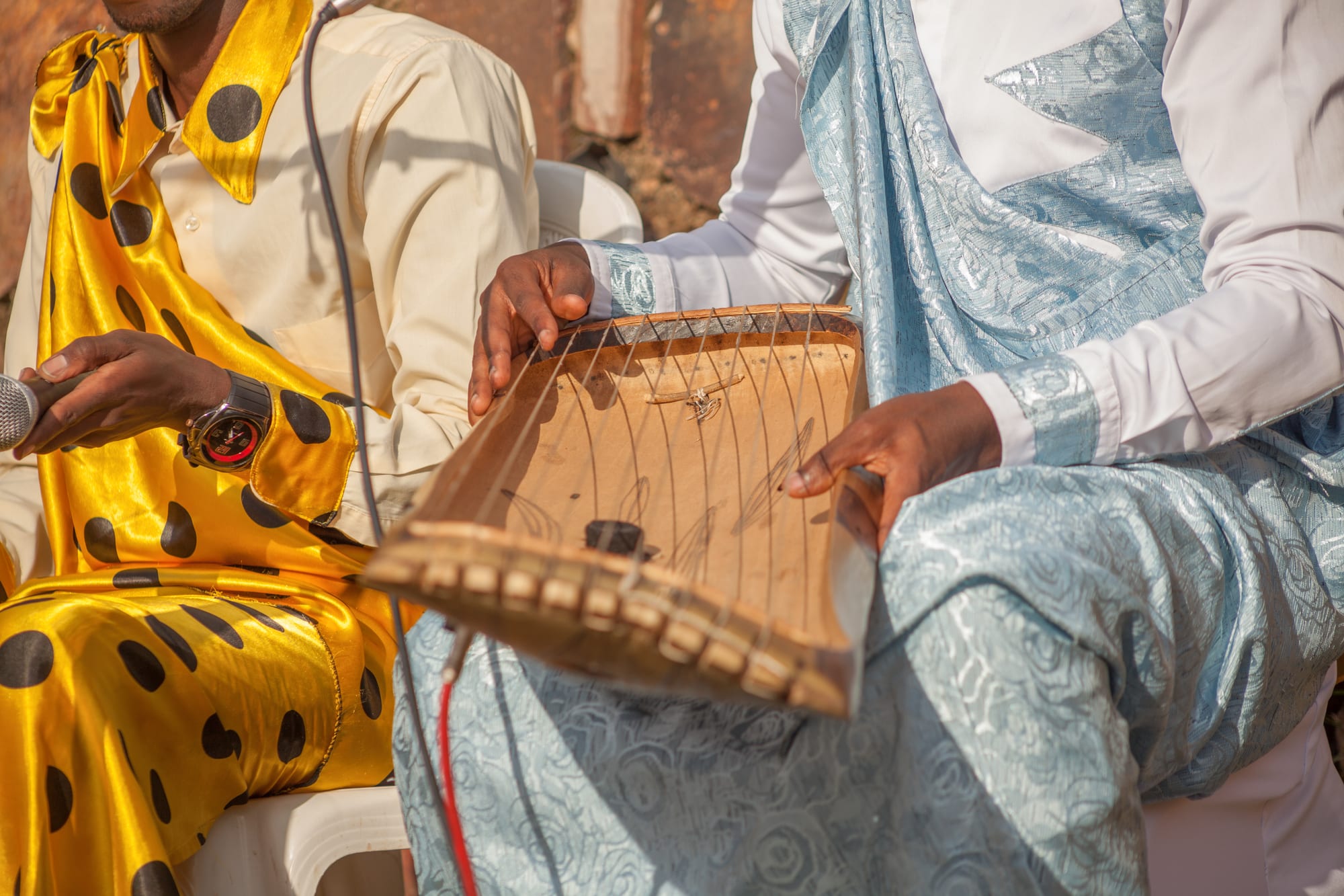 Close-up of a musician's hands plucking the strings of a flat wooden trough zither resting on his lap, beside another man in a yellow and black spotted outfit holding a microphone