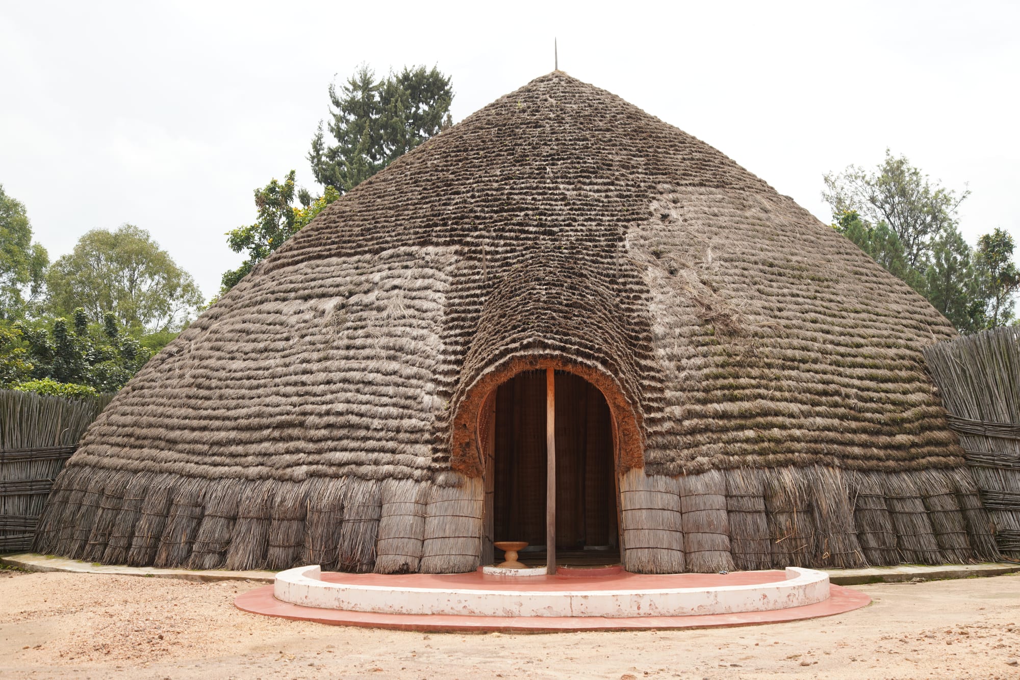A large dome-shaped building thatched from ground to peak with layered grass, with an arched entrance framed in reddish thatch and a curved step in front, surrounded by woven fences and trees