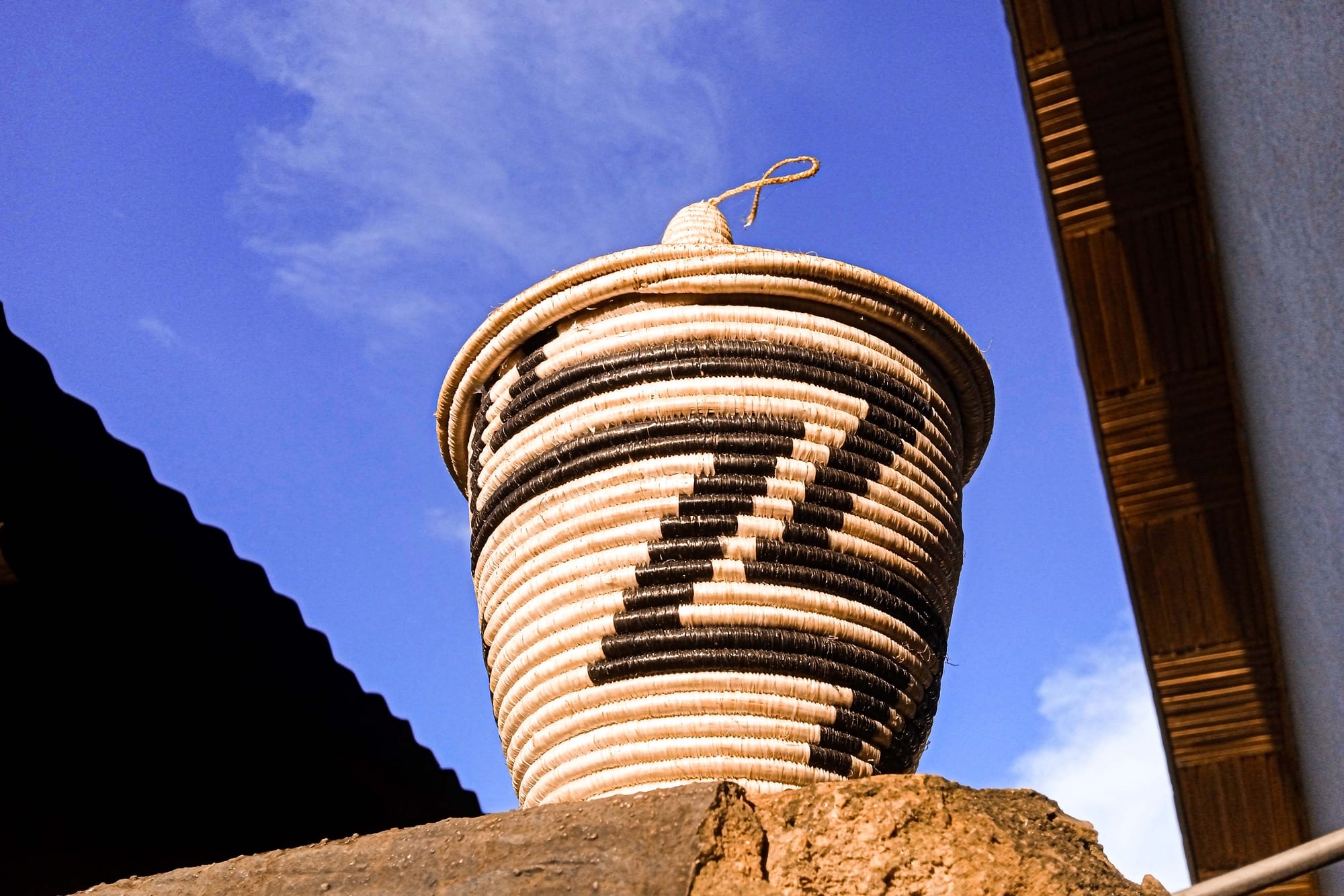 A tall coiled basket woven in cream and dark brown stripes with a zigzag pattern and a pointed lid with a small loop on top, standing on a stone wall against a blue sky