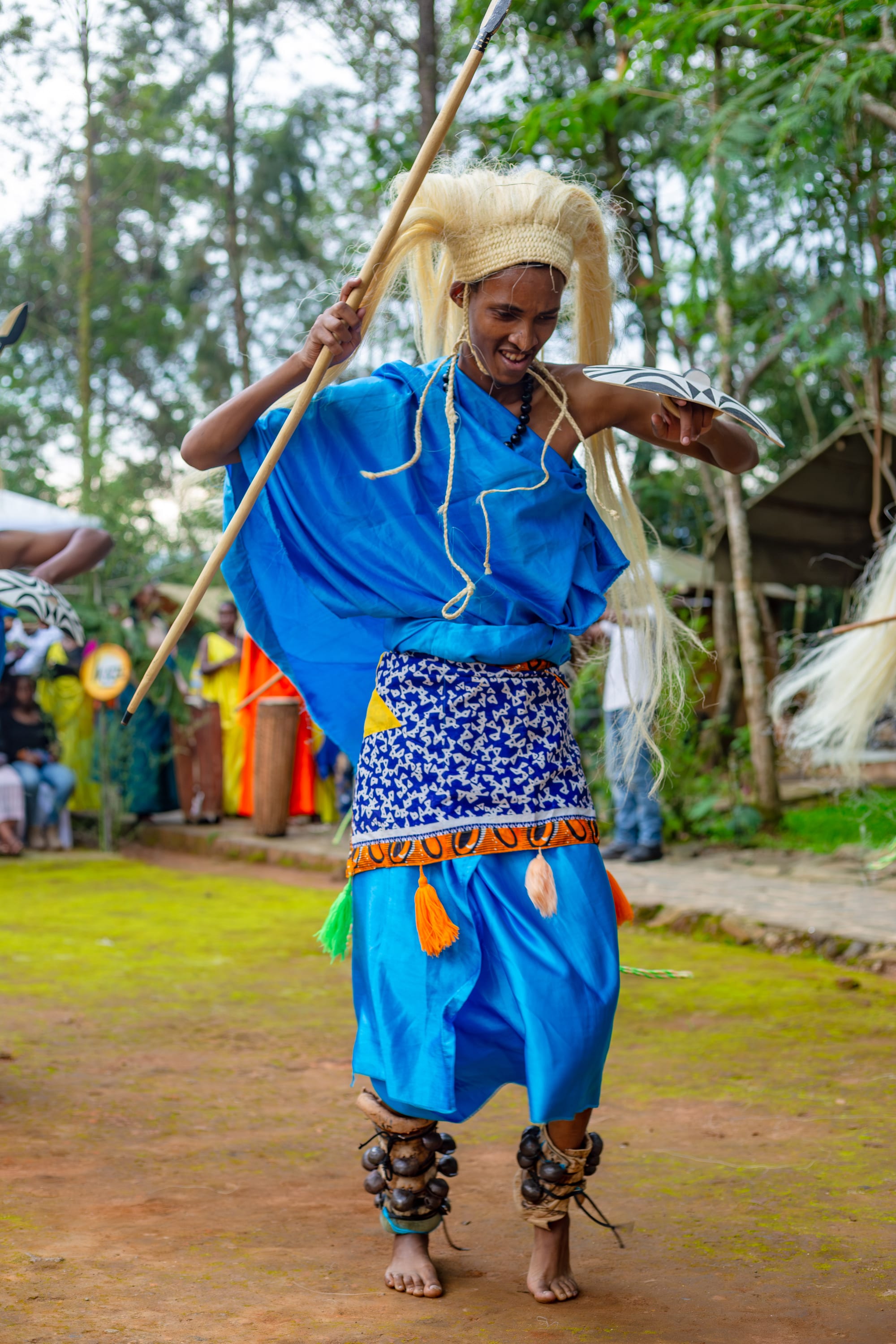 A dancer in a bright blue robe and patterned wrap, wearing a long pale headdress of fibre and bells on his ankles, leaps forward holding a spear and a small painted shield as spectators watch